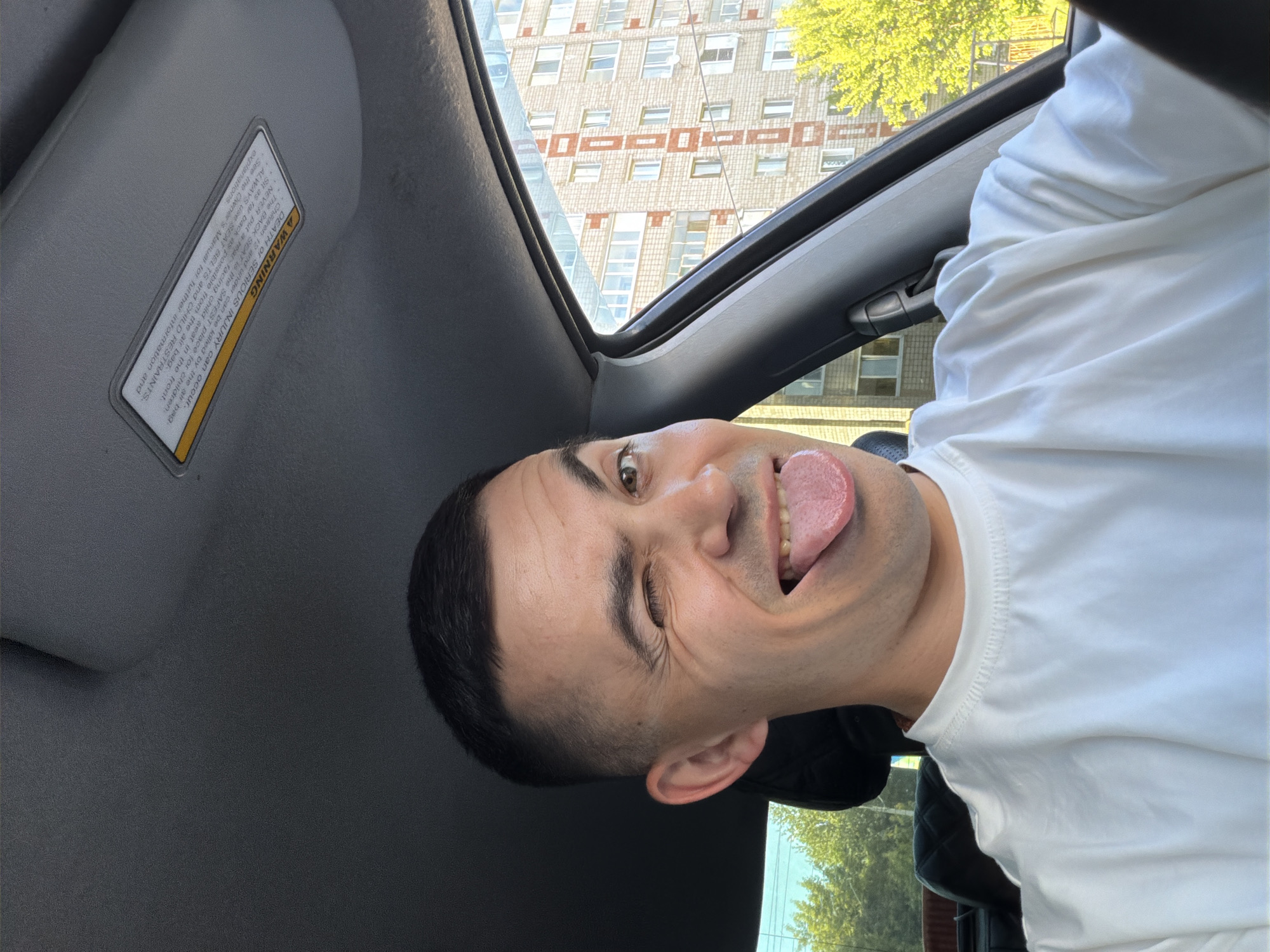 A man is captured in a playful selfie taken inside a car in Izhevsk, Russia. He has short, dark hair, light skin, and brown eyes, and is wearing a white short-sleeved t-shirt. He is seen winking with his right eye and sticking out his tongue, conveying a humorous and lighthearted emotion.
The perspective is from inside the car, looking upwards and outwards. A gray sun visor with a warning label is visible in the upper left, while the car's black interior trim frames the view of the outside world.
Beyond the car, a multi-story brick building with numerous windows and distinctive reddish-brown horizontal decorative bands is prominent in the background. Lush green foliage from trees is visible on both the right and upper left sides of the exterior view, suggesting a sunny daytime setting with clear weather.
The warning text on the car's visor reads: "A WARNING DEATH OF SERIOUS INJURY can occur. The BACK SEAT is the SAFEST place for children. Children 12 and under can be killed by the air bag. NEVER put a rear-facing child seat in the front. ALWAYS use SEAT BELTS and CHILD RESTRAINTS. Sit as far back as possible from the air bag. See the Owner's Manual for further information and explanations."