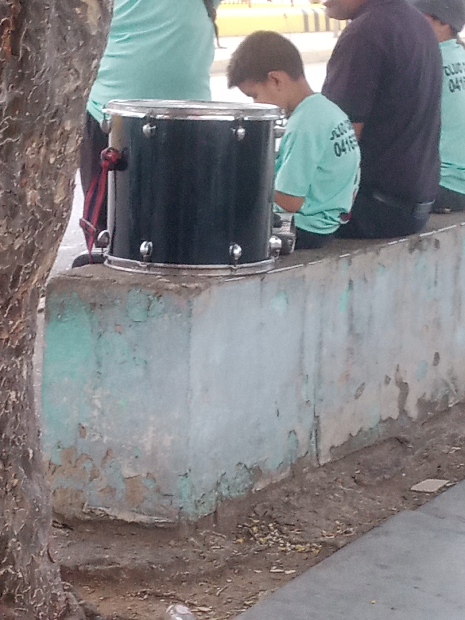 In Santa Ana, Venezuela, an outdoor scene depicts a group of people near a concrete structure. A young boy wearing a light blue t-shirt with white lettering, possibly a phone number, is seated beside a black drum. He is looking down, seemingly focused on something near the drum. To his right, an adult wearing a dark purple t-shirt is also seated, partially obscuring another individual in a light blue t-shirt. The drum, with silver hardware, rests on the concrete structure which shows signs of peeling paint, revealing a lighter surface underneath. A tree trunk is visible on the left side of the frame. The ground is covered in dirt and scattered debris.