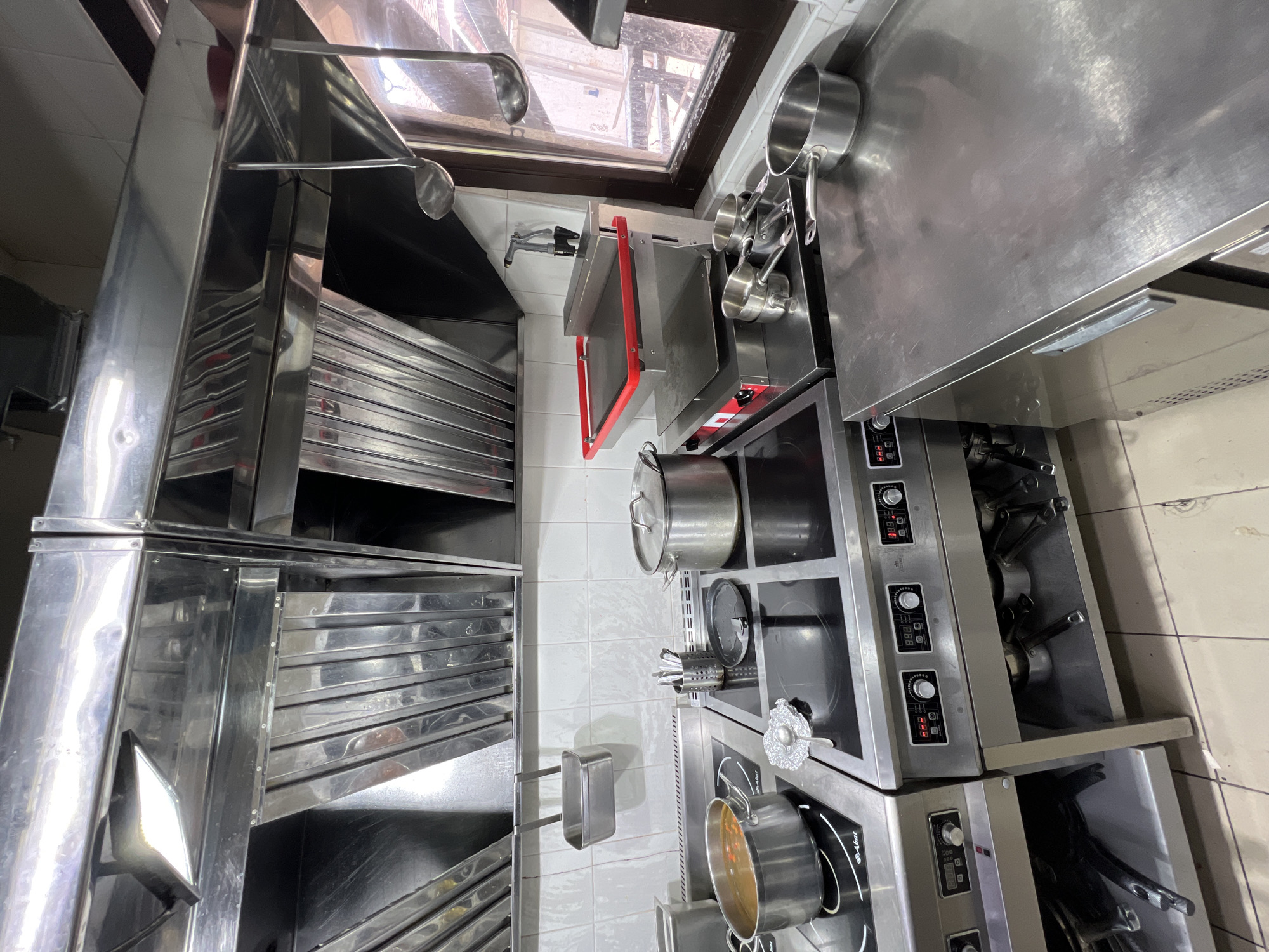 The image shows a commercial kitchen environment, likely in Plast univska, Russia. The scene is dominated by stainless steel surfaces and appliances. In the foreground, a stainless steel workbench is visible, with a handle on the left side. Behind it, a multi-burner induction cooktop is present, with various pots and utensils on its surface. One pot contains a liquid with some solid ingredients. To the right of the cooktop, a tiered cooking apparatus with a red-rimmed top shelf is set up. Further to the right, a window offers a view of an outdoor area with wooden structures. Above the cooking area, a stainless steel exhaust hood is installed, with ladles hanging from it. The walls are tiled with white square tiles. The overall impression is of a functional, busy kitchen, though no people are visible in the frame.