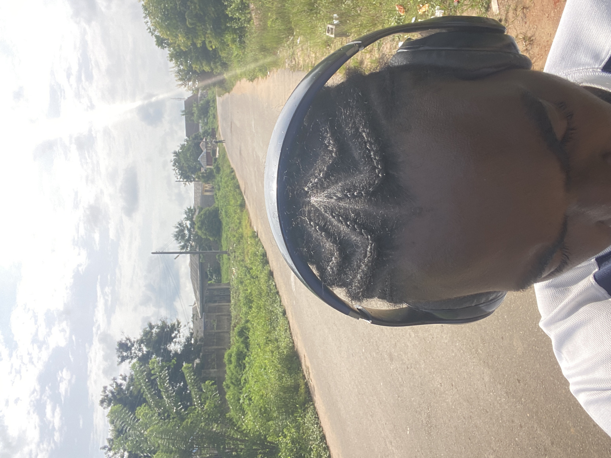 The image shows a close-up of a person's head and shoulders, wearing headphones. The person has their hair braided in a star-like pattern. They are wearing a white shirt with blue accents. The background is an outdoor scene with a paved road stretching into the distance. There are buildings and lush green vegetation on either side of the road, under a cloudy sky with bright sunbeams. In the distance, a lone figure is walking on the road. The scene appears to be a residential area in Oniyangi, Nigeria, possibly during daytime.
