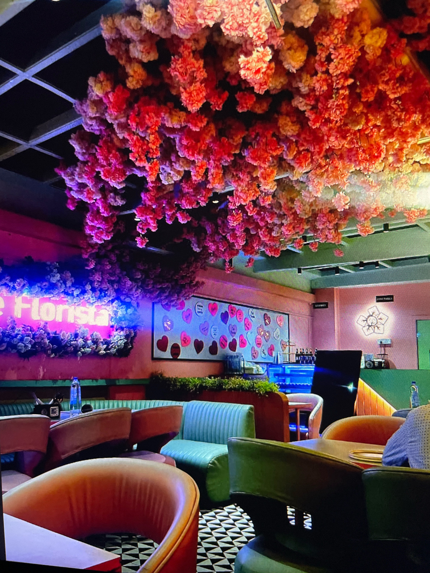 The image depicts the interior of a cafe or restaurant, possibly named "The Florista" as suggested by a neon sign. The space is decorated with an abundance of pink and red floral arrangements hanging from the ceiling and along the walls. The seating area features a mix of plush chairs and booths in various colors, including teal, orange, and green. A bar area with blue lighting is visible in the background, with bottles of alcohol on display. A framed artwork featuring hearts is mounted on a pink wall. The floor has a distinctive black and white geometric pattern. The overall ambiance appears to be vibrant and lively, with soft lighting enhancing the colorful decor. There are no people clearly visible in the foreground, though a person is partially seen in the background on the right. The location is specified as Gankar, India.