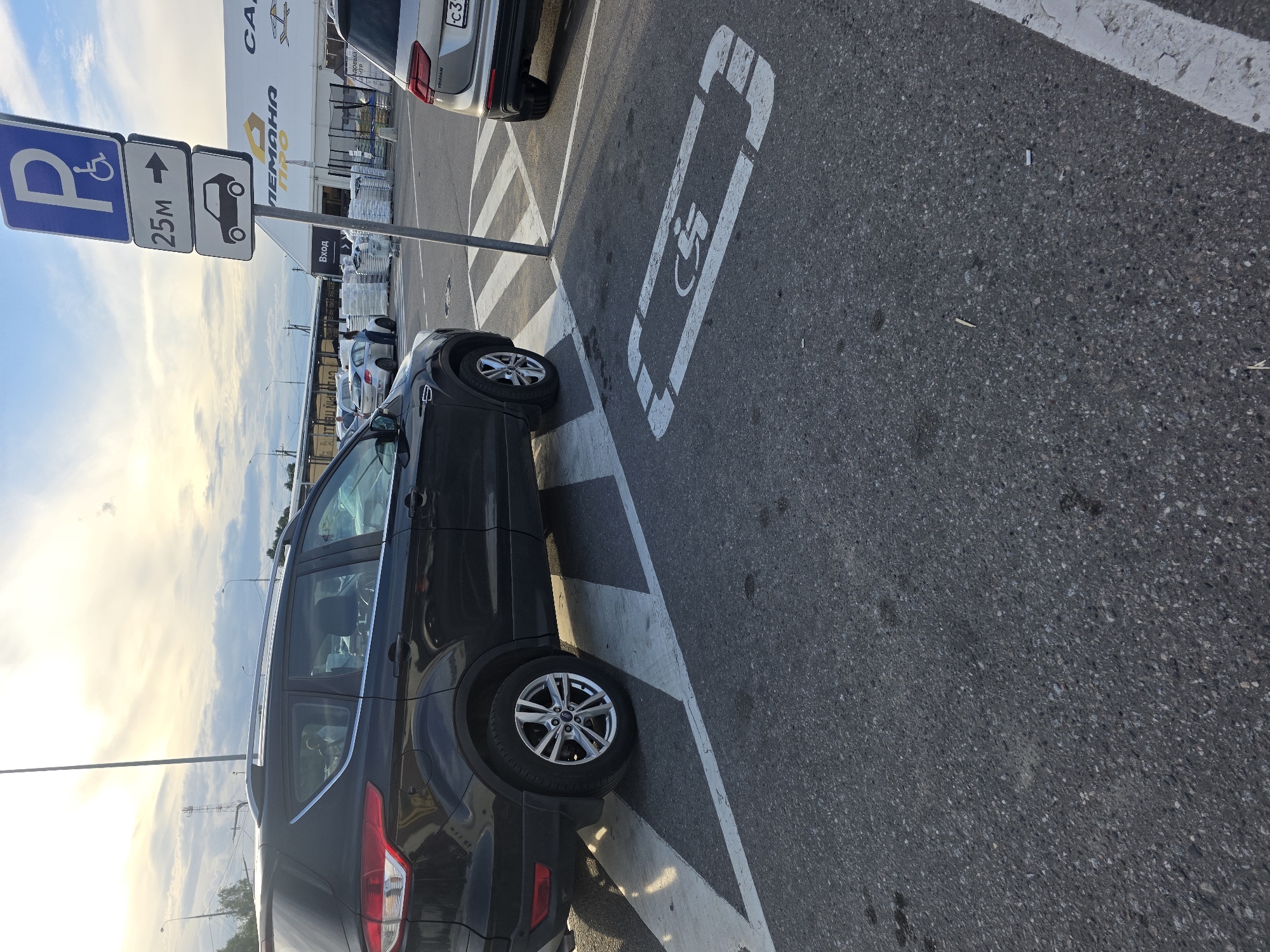 A black SUV is parked in a parking lot, its tires on a white-lined, handicapped parking space. The space is marked with a white wheelchair symbol painted on the asphalt. A signpost stands next to the parking spot, displaying three signs. The top sign is blue with a white wheelchair symbol and a white "P". Below it is a rectangular sign with a white arrow pointing to the right and the text "25m". The bottom sign is a white rectangle with a silhouette of a car and a white arrow pointing to the right. In the background, a modern building with large windows and some retail signage is visible. The building has "РЕМАНа" and "СА" written on it in large letters. Other cars are parked in the vicinity, and a person is visible near one of them. The sky is partly cloudy with a bright, hazy sun, suggesting it is either early morning or late afternoon. The scene appears to be a public parking area, possibly outside a shopping center or commercial establishment.
