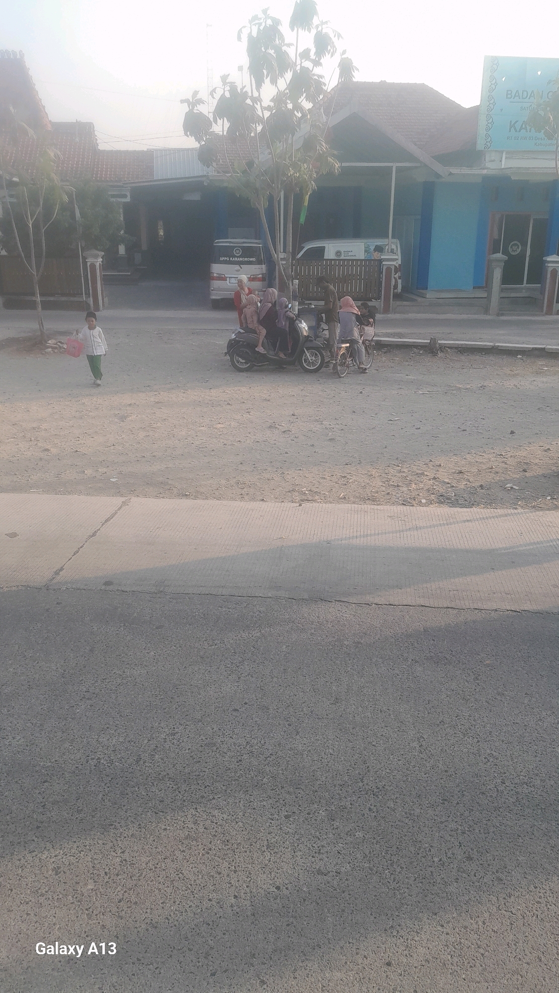 In Ngelo, Indonesia, at what appears to be the entrance to a building with a blue facade and a red-tiled roof, a group of individuals are gathered near a scooter and a bicycle. A child in a white shirt and green pants walks across a dirt lot in the foreground, carrying a pink object. Behind the child, a black scooter is parked, with several people, including women and children, on and around it. One woman, wearing a pink hijab, sits behind the driver. To the right of the scooter, a bicycle is parked, with a person in a light brown hijab standing next to it. Further back, a white van with "SPPO KARANROWO" written on its side is parked in front of the building. Another white vehicle with a logo is also visible. The building has a sign that reads "BADAN C" and other text that is difficult to decipher. The scene is illuminated by natural light, casting shadows on the ground.