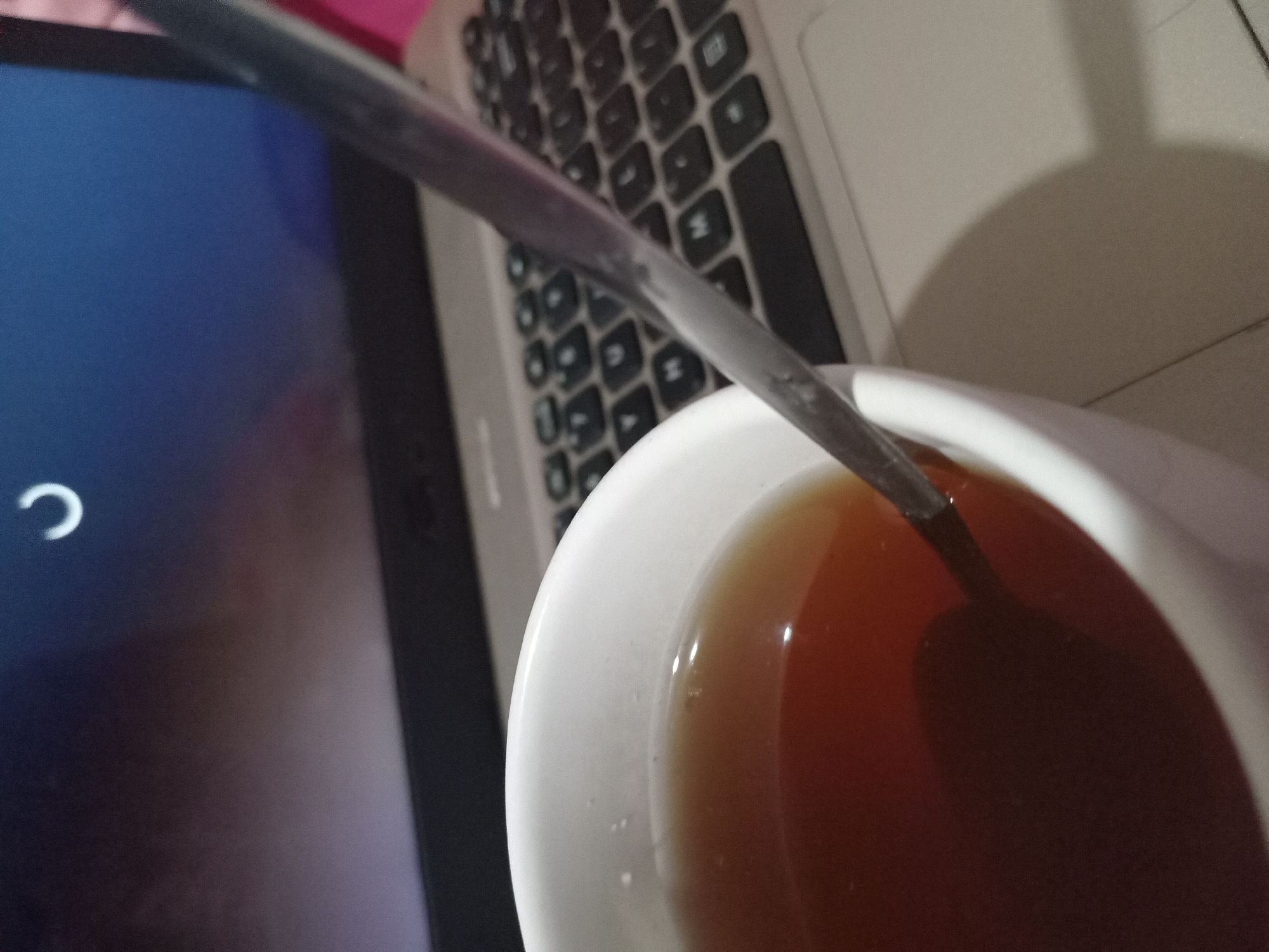 The image depicts a close-up of a white mug filled with amber-colored liquid, likely tea, with a silver spoon resting inside. The mug is positioned on a laptop keyboard, suggesting a work or study setting. In the background, the laptop screen displays a loading icon, a white curved line against a dark blue and purplish gradient. The overall scene is dimly lit, with the light source appearing to be the laptop screen. The image does not provide any specific geographical information about Claket, Indonesia, nor does it show any individuals or explicit actions beyond the presence of the objects.