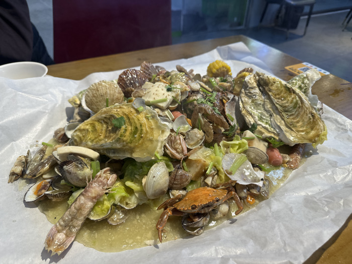 A large communal seafood platter is centrally displayed on a wooden table, served directly on a sheet of white paper. The dish consists of a variety of shellfish and other ingredients immersed in a pale, yellowish-green sauce or broth. Prominent items include multiple oysters, numerous clams of various types, at least two scallops, one reddish-orange crab, one large mantis shrimp, and sections of corn on the cob. Green leafy vegetables, possibly cabbage, and chopped herbs like cilantro are interspersed throughout the seafood. In the background, to the left, a person wearing a dark garment is partially visible, seated at the table, with a small white bowl positioned nearby. The wider background suggests an indoor dining establishment with dark walls, a reddish-brown panel, and blurred furniture. This scene is located in Huangdao District, China.