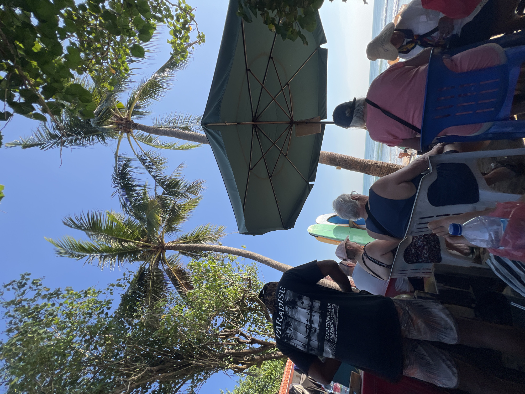 In Kuta, Indonesia, a group of people are seated under a large green umbrella near the beach. A man in a black t-shirt that reads "PARADISE" and "GOOD THINGS ARE FUCKING COMING" stands to the left, facing away from the camera. In front of him, several people, including a woman in a pink shirt and a man in a blue sleeveless shirt, are seated in chairs, facing the ocean. Surfboards are visible behind the seated individuals, and palm trees and lush green foliage frame the scene against a clear blue sky. A plastic bottle of water sits on a table in the foreground.