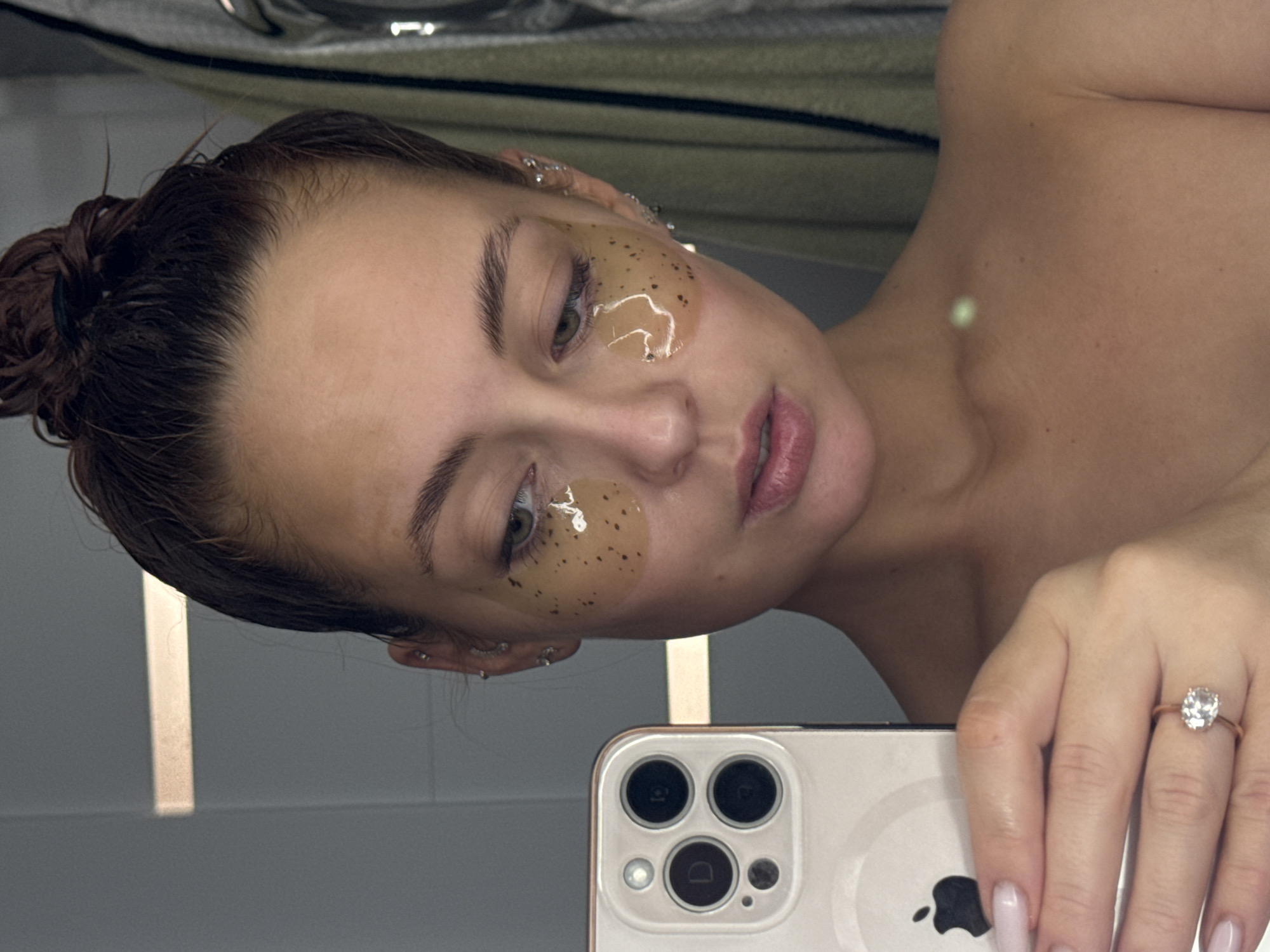 A female individual is depicted taking a mirror selfie in an indoor setting, likely a bathroom. She has dark hair pulled back into a wet bun and is wearing translucent under-eye patches containing dark particulates beneath both eyes. Multiple piercings are visible on her left ear. Her right hand, adorned with a diamond ring on the left ring finger, holds a light-colored iPhone (identifiable by the Apple logo and triple-lens camera module) in the lower left portion of the frame. Her gaze is directed downwards towards the phone. The background consists of light grey tiled walls, with a horizontal light fixture reflected in the upper left. On the right, partially visible, are hanging textiles, possibly towels or garments, in shades of green and grey. No visual identifiers within the image provide information regarding its specific location in Chaikovsky, Russia, or any other city.