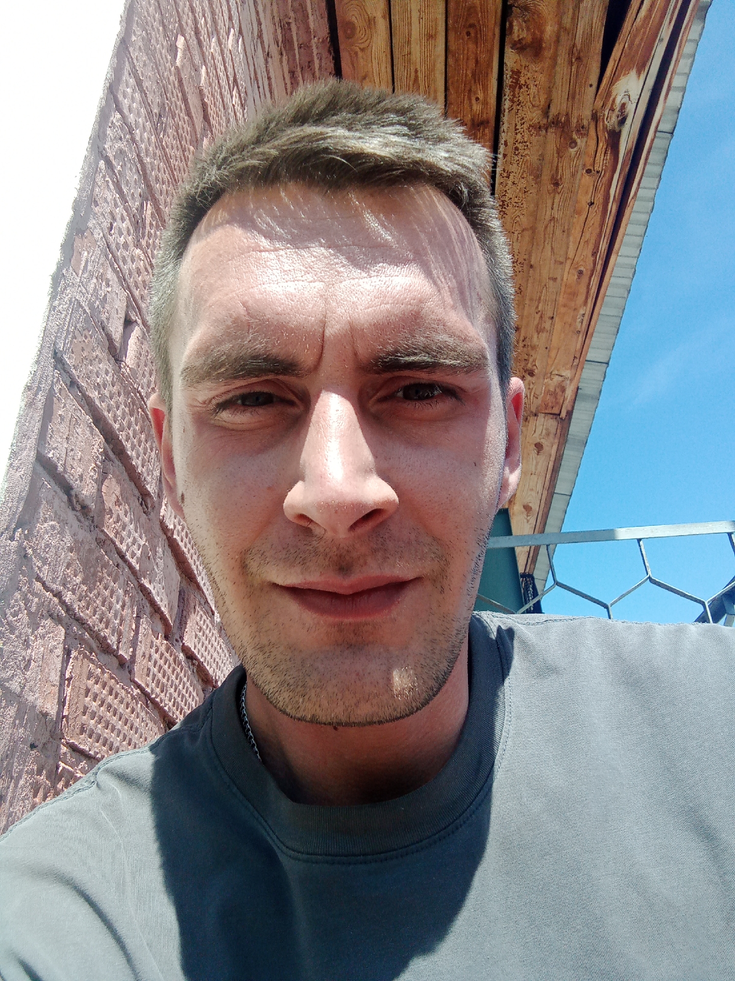 The image is a close-up selfie of a fair-skinned man with short, dark hair and stubble. He is wearing a gray T-shirt. Behind him are a brick wall on the left and wooden planks with a blue sky visible above on the right. A metal fence is visible at the bottom right. The location is Chaikovsky, Russia.