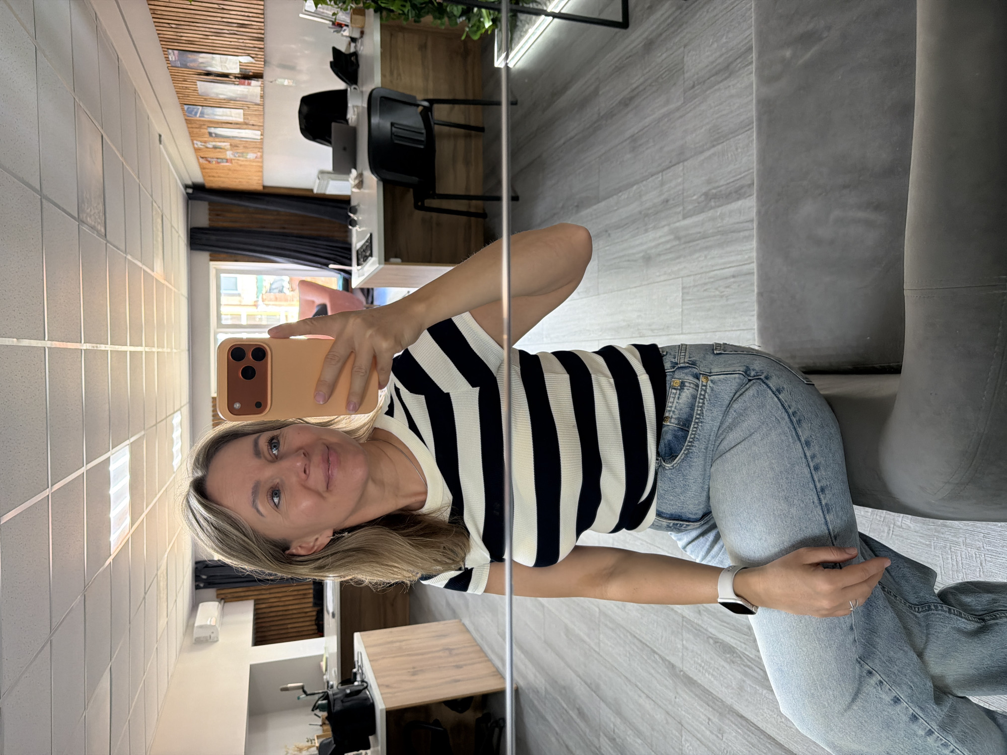 A woman is taking a mirror selfie in an office setting. She is wearing a black and white striped short-sleeved shirt and light blue jeans. She has blonde hair and is smiling. In the reflection, we can see office desks with computers, chairs, and decor. The floor is made of grey wood-like planks, and the walls are decorated with wooden slats and posters. The office appears to be well-lit with fluorescent ceiling lights and natural light coming from windows. The city of Chaikovsky, Russia, is not discernible from the image.