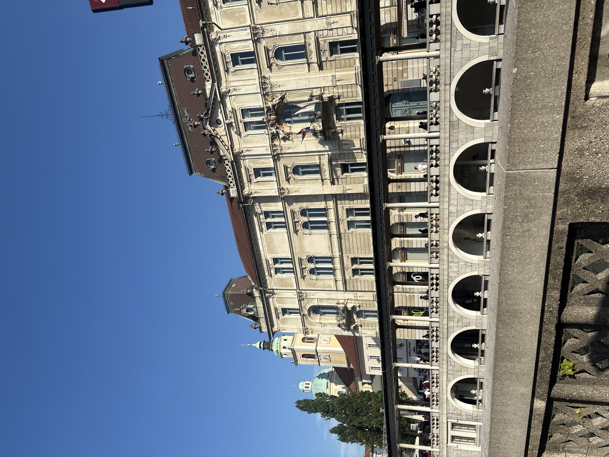 The image depicts a classic architectural scene in a city square. A grand, multi-story building with ornate detailing and a reddish-brown roof dominates the right side of the frame. It features numerous windows, balconies, and decorative elements. To the left of this building, a church tower with a green dome and clock face rises, followed by another building with a similar green dome. In the foreground, a colonnade with arches runs along the base of the main building, with people strolling and seated at tables, suggesting a public space like a cafe or walkway. Below the colonnade is a stone wall with decorative patterns, and further in the foreground, a rougher stone embankment overlooks what appears to be water, with reflections visible. The sky is a clear, vibrant blue. The overall impression is one of historical significance and urban activity under a bright sky.