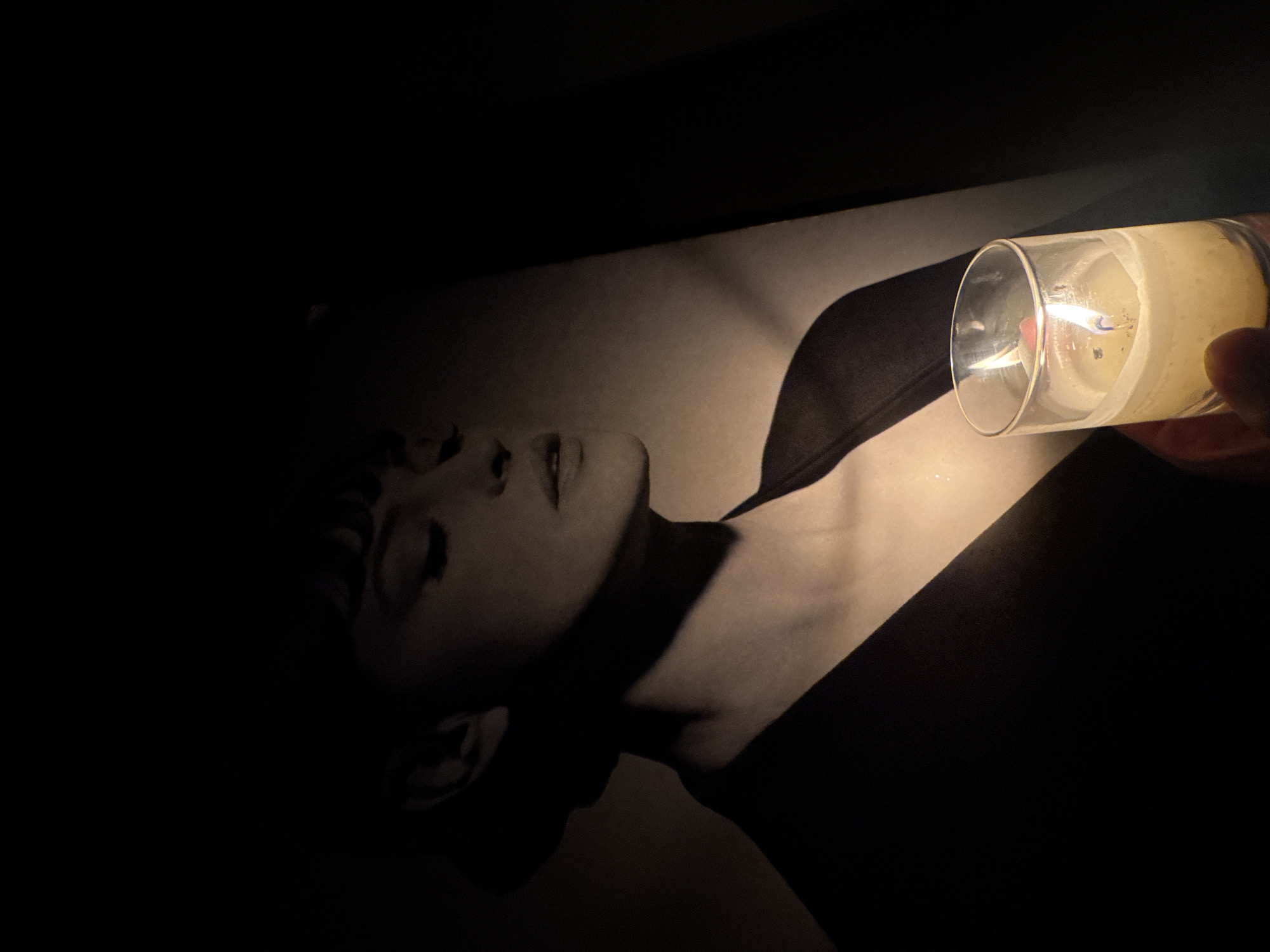 A close-up, high-angle shot depicts a black-and-white photograph of a person's face and upper torso illuminated by the light of a candle held in a clear glass. The photograph lies on a dark surface, with the left side of the image obscured in shadow. The candle is positioned on the right side, its flame visible. The photograph appears to show a person lying down, with their face turned upward. No information suggests that this image was taken in Sofia, Bulgaria.