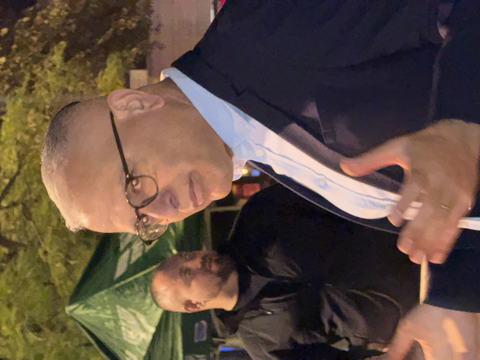 A medium shot captures two men, both bald. One wears glasses and a collared shirt underneath a dark suit jacket. His hand is visible with a ring on his finger. The other man, out of focus, is dressed in black. A green tent is slightly visible in the background. The photo appears to be taken outdoors at night.