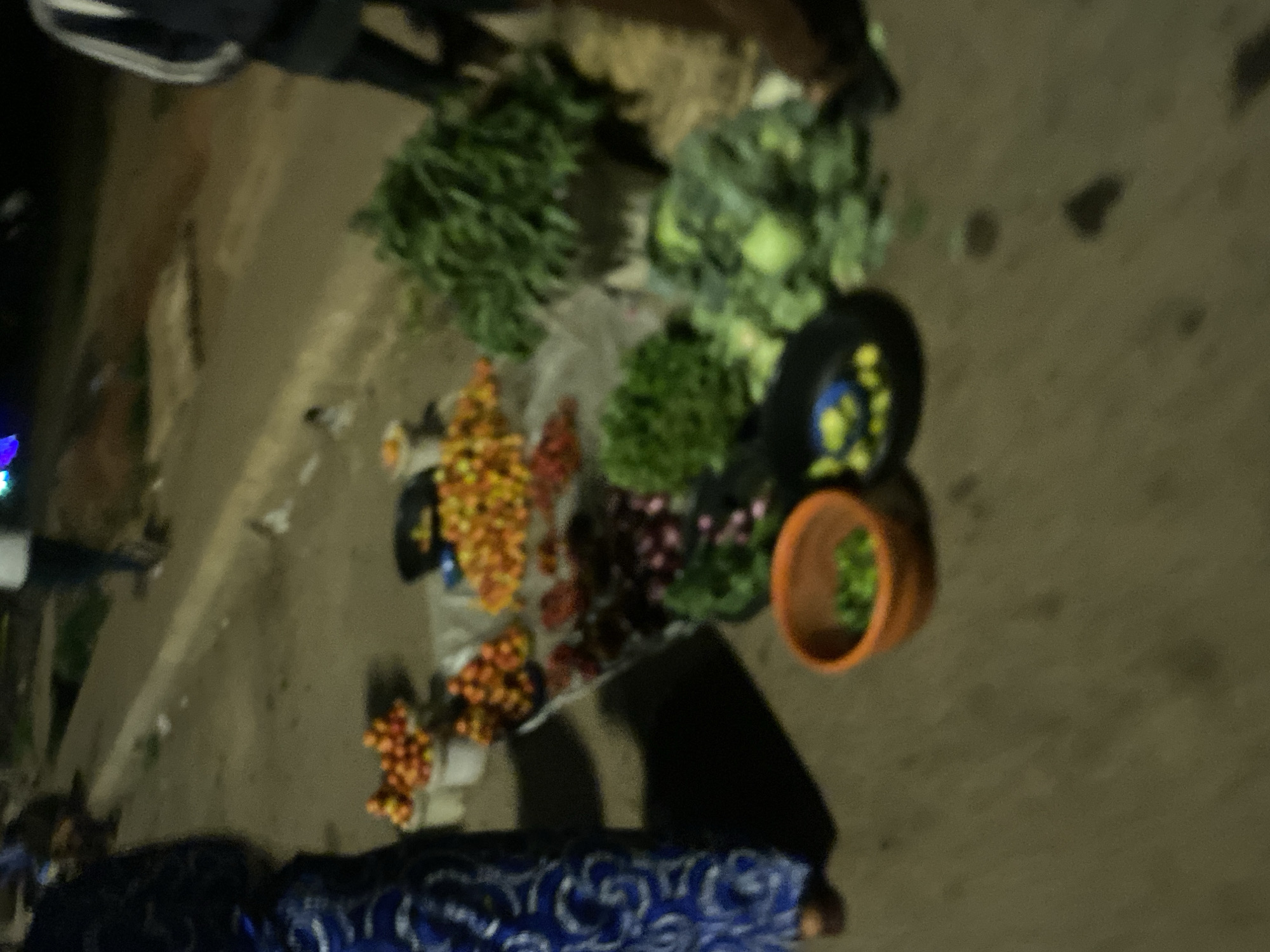 The image depicts a street-level view of a produce stall in Bukuru, Nigeria, at dusk or night. A person wearing a blue and white patterned garment stands to the left, partially obscuring the scene. The stall displays a variety of fresh produce, including red tomatoes in white buckets, an assortment of orange-colored fruits, and green leafy vegetables. There are also items that appear to be eggplants or similar purple vegetables. Two shallow bowls are visible; one orange bowl contains green leafy items, and a black bowl holds round, yellow objects. The ground is paved with asphalt. In the background, blurred figures of other people and a paved sidewalk suggest an urban or semi-urban environment. The lighting is dim, indicative of evening or night conditions.