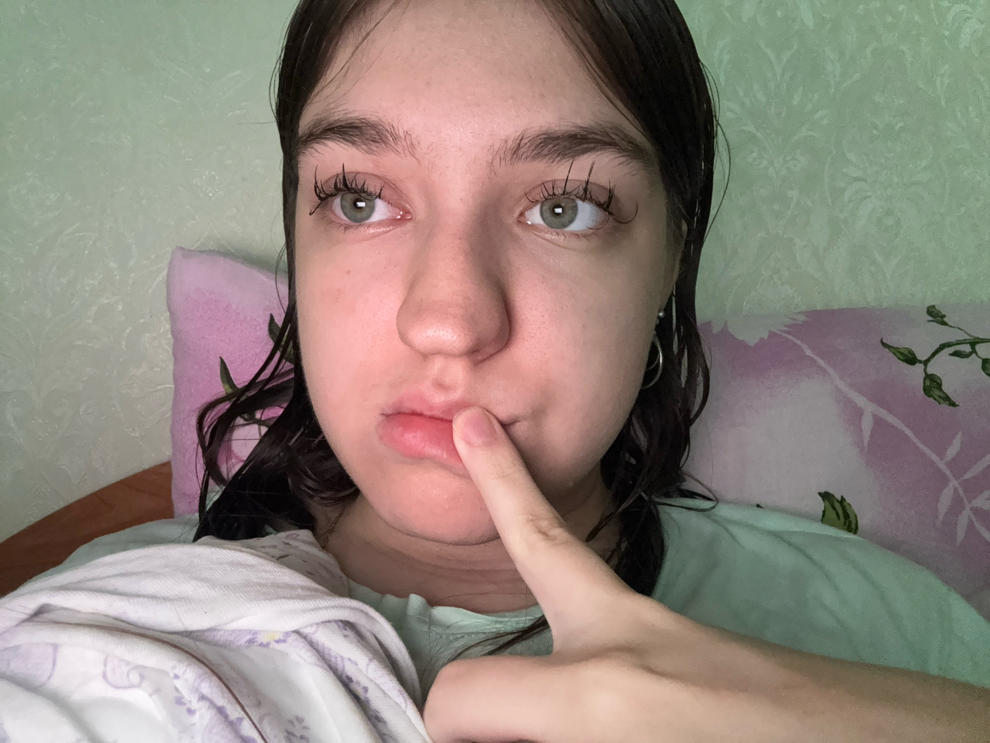 A close-up, head-and-shoulders portrait shows a young person with dark, wet hair, looking to their left. They have fair skin, prominent eyelashes, and light green eyes. Their right index finger is touching their lower lip. The person is wearing a light blue t-shirt. Behind them, a light green textured wallpaper is visible, with a pink and white floral patterned pillow or blanket to their left and a similar patterned blanket or sheet in the foreground. The overall impression is of a personal, indoor setting, possibly a bedroom, likely in Zavolzhye, Russia, given the context.