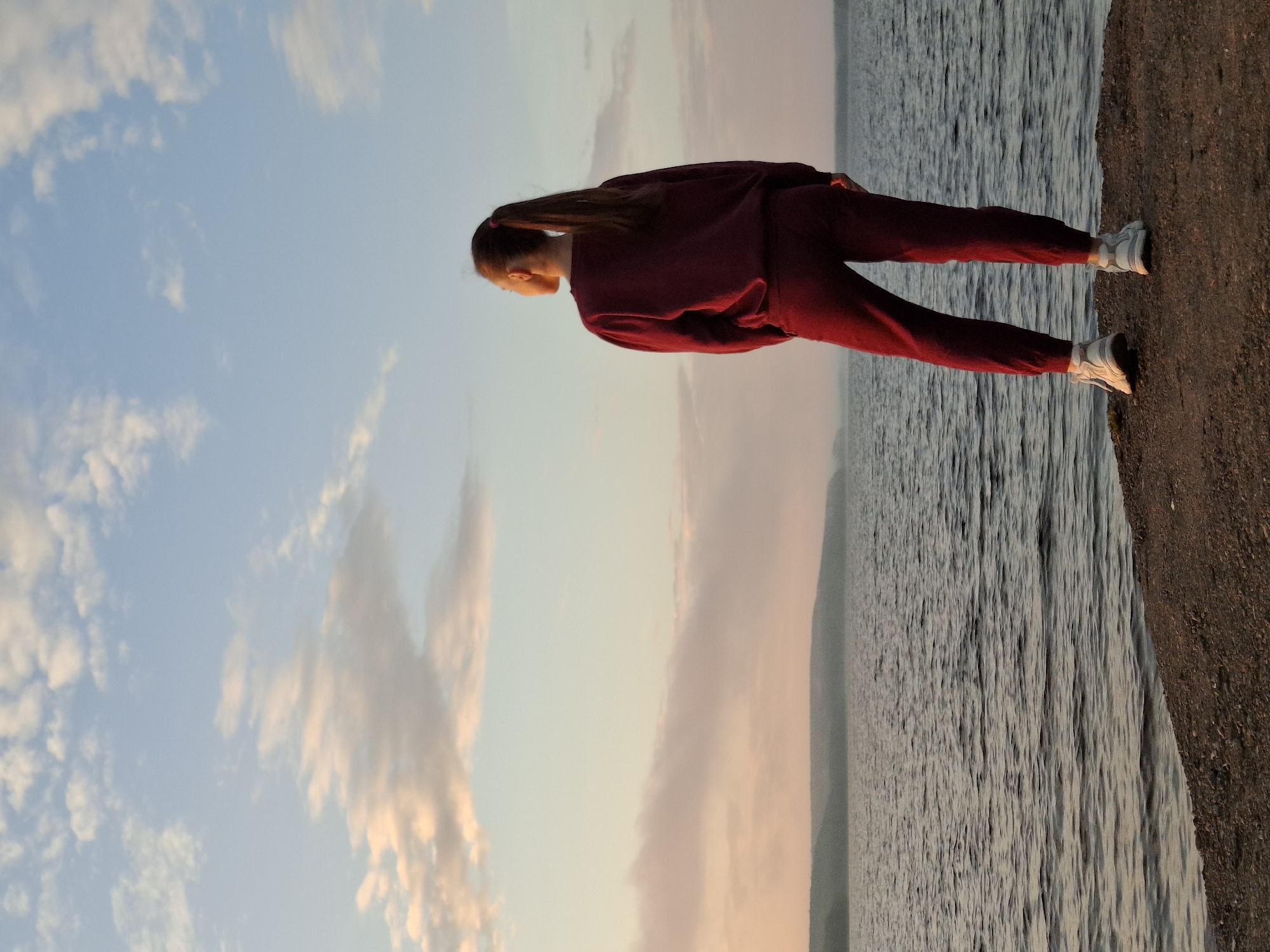 A lone woman stands on a rocky shore, facing a body of water and a distant, hazy shoreline. The sky above is a gradient of pale blue and soft pinks, with scattered clouds catching the golden light of either sunrise or sunset. The woman is wearing a dark red tracksuit and white athletic shoes. Her back is to the camera, and her hair is pulled back in a ponytail. The water is calm, with gentle ripples reflecting the sky. The overall atmosphere is peaceful and contemplative. The scene is likely located near Chaikovsky, Russia, given the characteristic landscape of the Kama River basin.