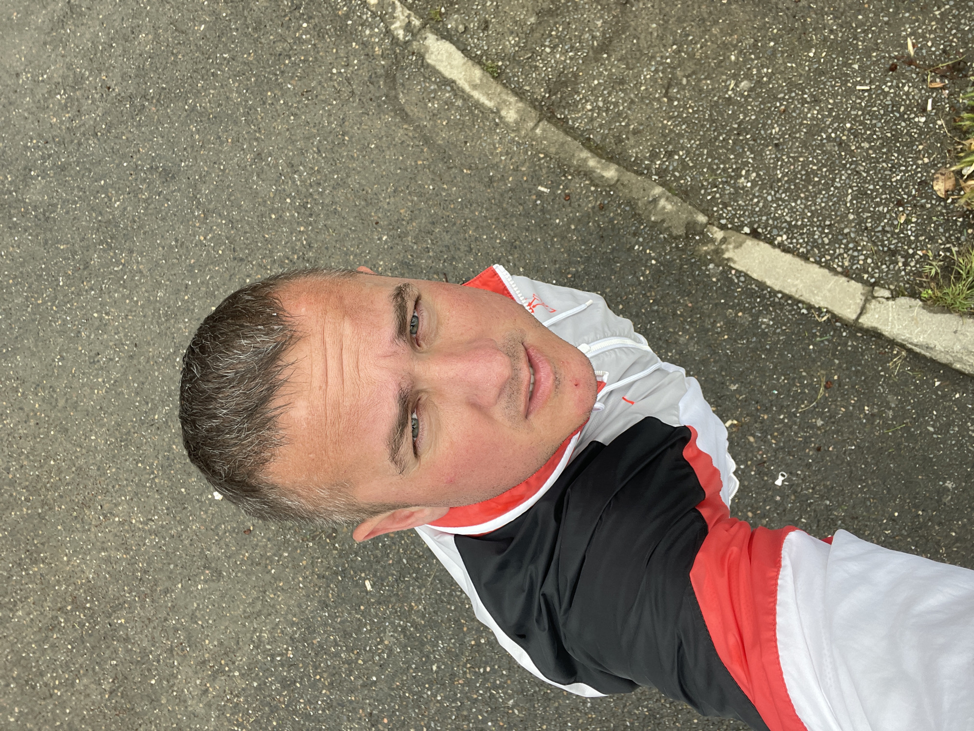 The image shows a medium-shot of a man with short gray hair and fair skin, wearing a track jacket that is black, red, and white. He is looking at the camera. The background is asphalt. It is impossible to tell if the image was taken in the city of Sochi, Russia, with the available information.