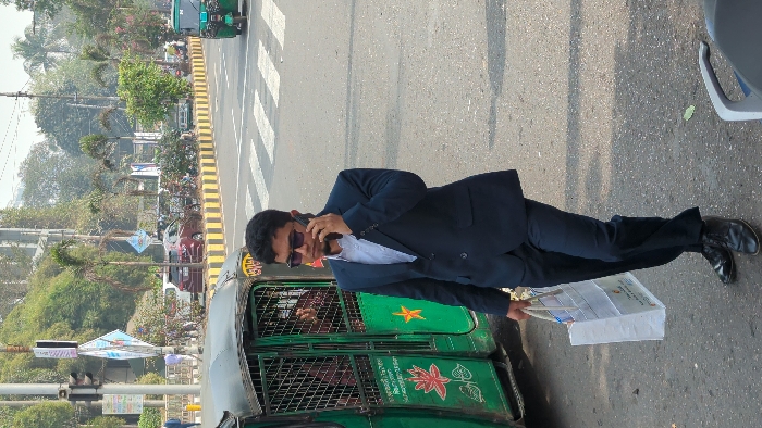 A man in a dark suit stands next to a green auto-rickshaw on a street. He holds a white bag or package in one hand and is talking on a cell phone. The street has crosswalk markings and a yellow-striped divider. Trees and other vehicles are visible in the background, suggesting an urban environment.