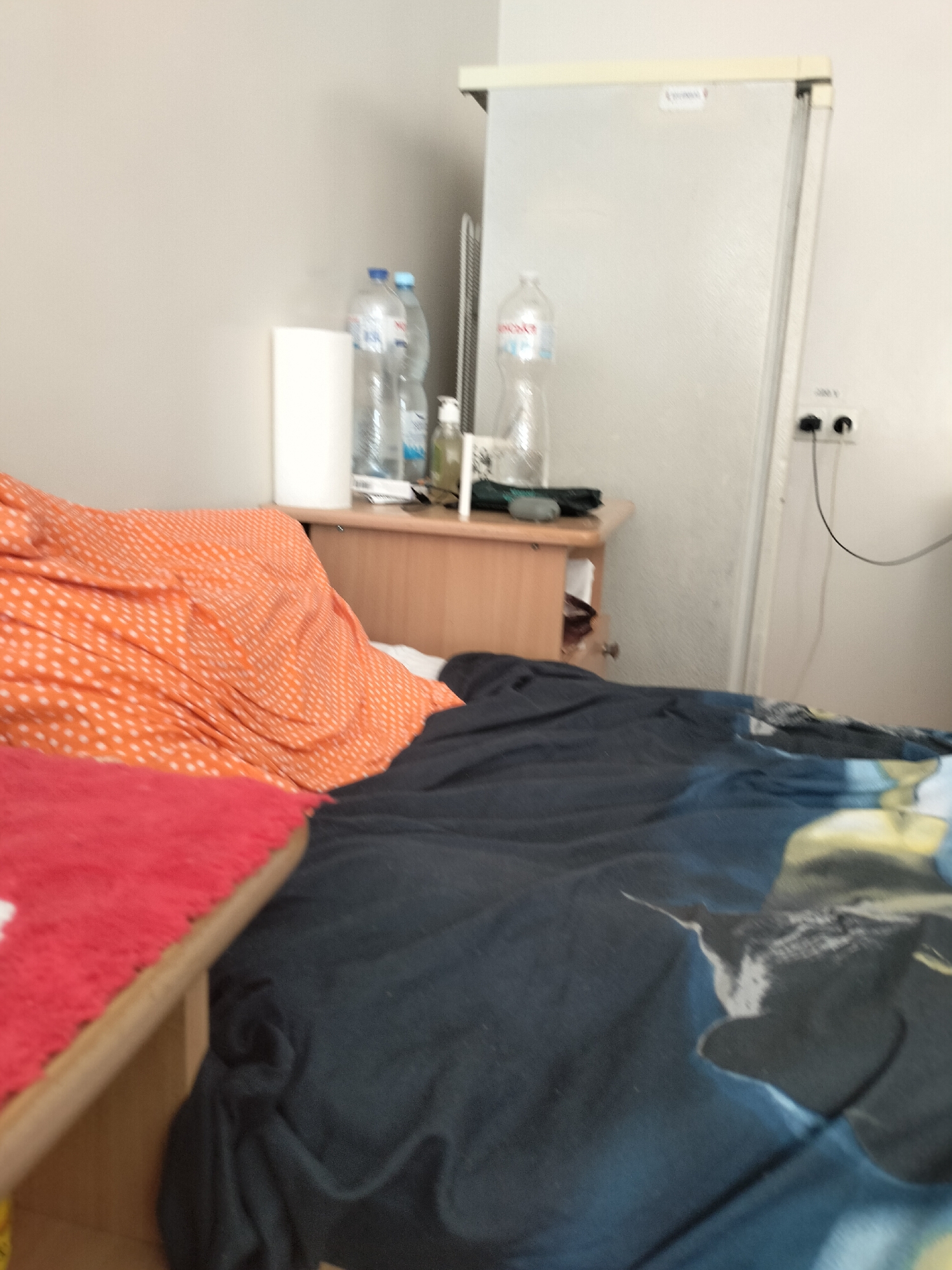 The image shows a messy bedroom. There is a bed with an orange and white polka dot blanket and a dark blue blanket with a graphic print. There is also a red mat on a wooden surface. In the background, there is a light-colored refrigerator, a wooden cabinet with some bottles and a dispenser on top, and a white cylindrical object on the cabinet. There are electrical outlets and cords on the wall next to the refrigerator. The room appears to be in a state of disarray.