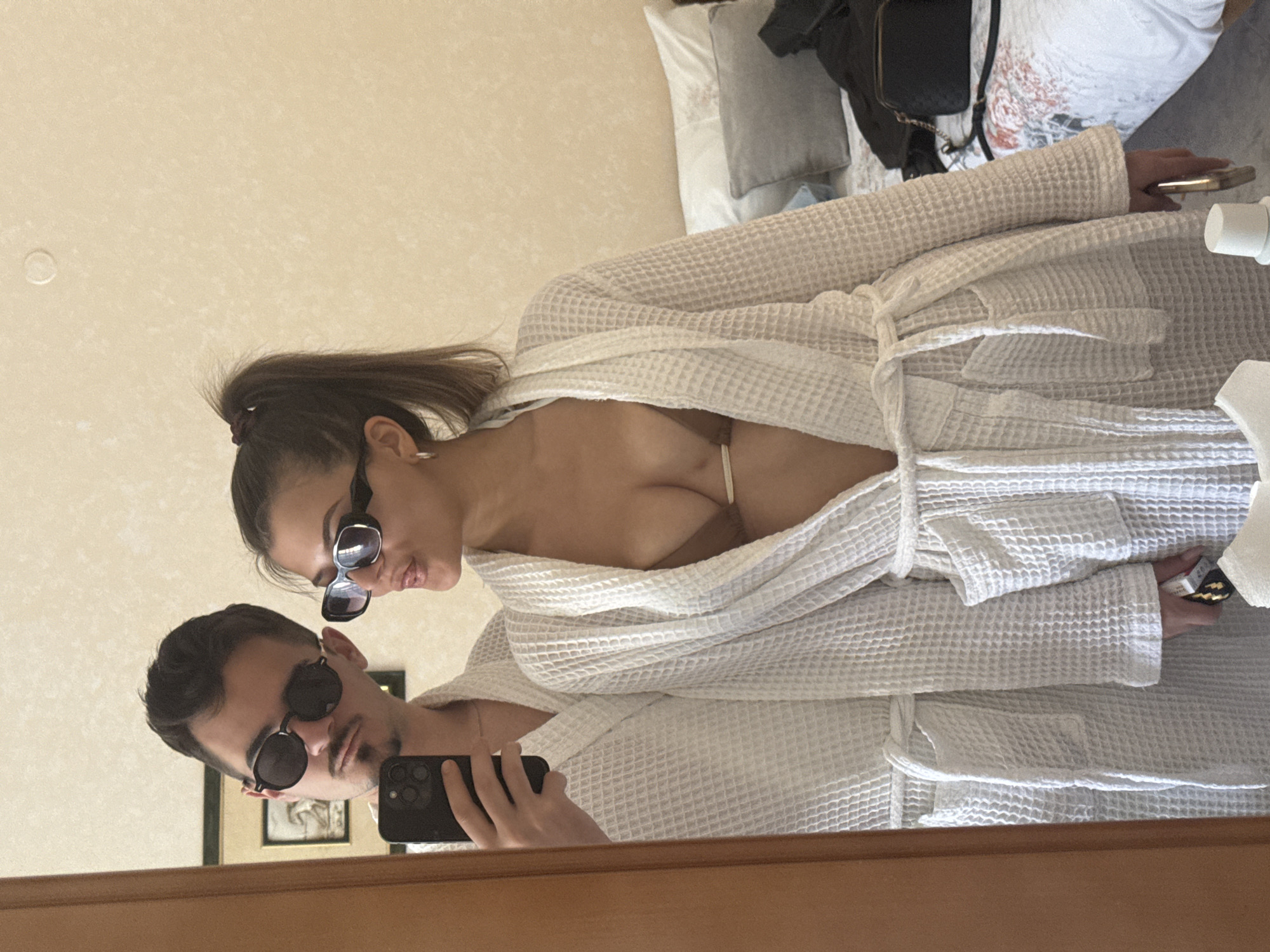 A mirror selfie taken inside a room shows a man and a woman wearing sunglasses and white waffle-weave bathrobes. The woman has her hair in a high ponytail. A portion of a bed with white linens, pillows, and a black bag is visible in the background. A framed picture hangs on the wall behind the man. The location appears to be indoors, possibly a hotel or home, but the specific city or region of Marchevo, Bulgaria, cannot be determined from the image alone.