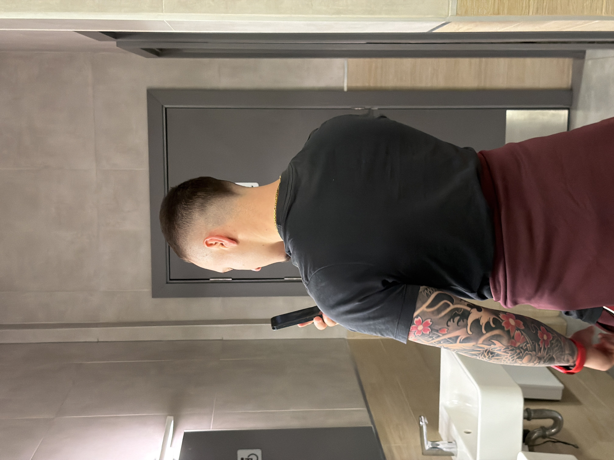 A man is standing in a restroom, looking at his phone. He has short hair and a black shirt, with a maroon garment around his shoulders. The man has a full tattoo sleeve on his left arm. The restroom has light grey tile walls, a grey door with a silver frame, and a white sink. The location is Plovdiv, Bulgaria.