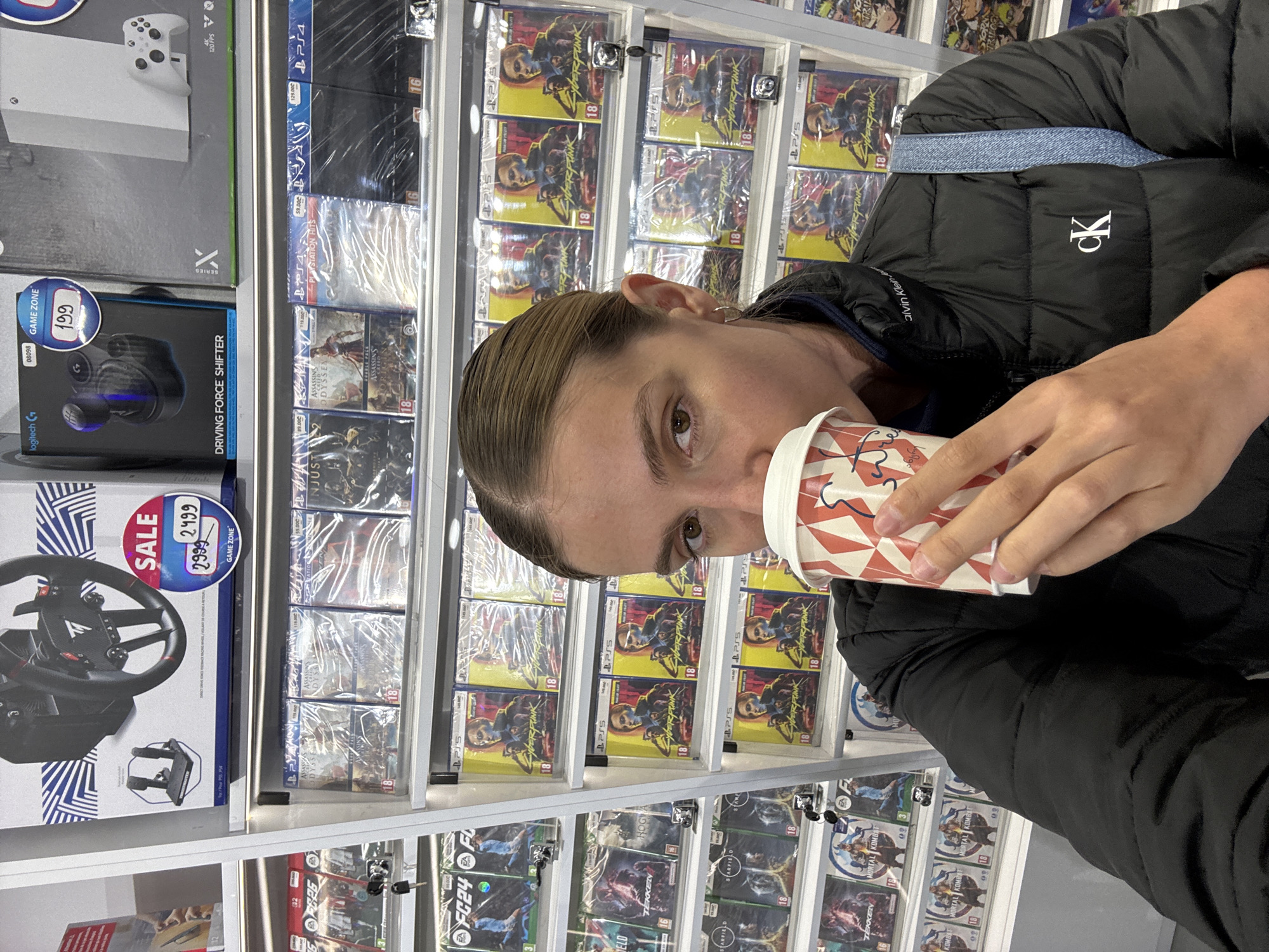 A person in a black Calvin Klein puffer jacket is seen holding a disposable cup. The background is a retail setting, with shelves stocked with video games, including "Cyberpunk 2077", "FC24", "Hogwarts Legacy", "Tekken", and "Mortal Kombat". There is also a video game console, driving wheel, and shifter on sale. The location is the GAME ZONE store in the city of Tbilisi, Georgia.