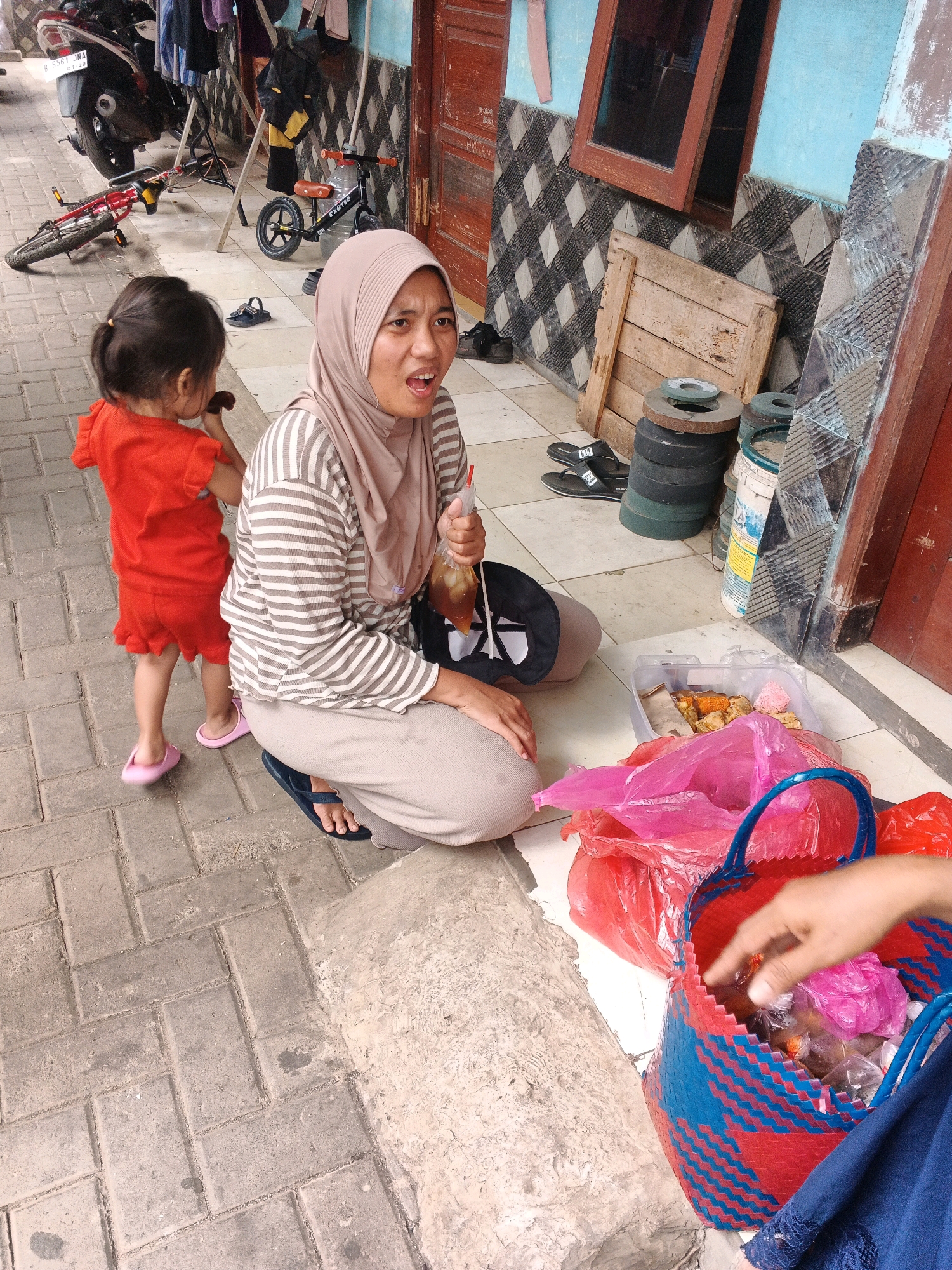 In the city of Kadu, Indonesia, an outdoor scene unfolds with a mother and child. The mother, wearing a hijab and a striped long-sleeved shirt, kneels on the ground, holding a plastic bag with a drink. Her mouth is open as if speaking. Beside her, a young child in an orange outfit stands with their back to the camera, facing away from the mother. They appear to be eating something.
The ground is paved with light gray interlocking bricks. To the right of the mother and child, various objects are scattered. A red and blue woven basket sits on the ground, filled with items wrapped in plastic. Several plastic bags, some pink and some red, are also present, along with a clear plastic container holding food. In the background, a wooden crate and stacks of what appear to be circular weights or grinding stones are visible. A small bicycle with a red frame lies on its side to the left, and another bicycle is parked upright. A motorcycle is partially visible on the far left. The wall behind the subjects is painted blue and features a checkered tile pattern. A wooden door is present to the right. The lighting suggests it is daytime.