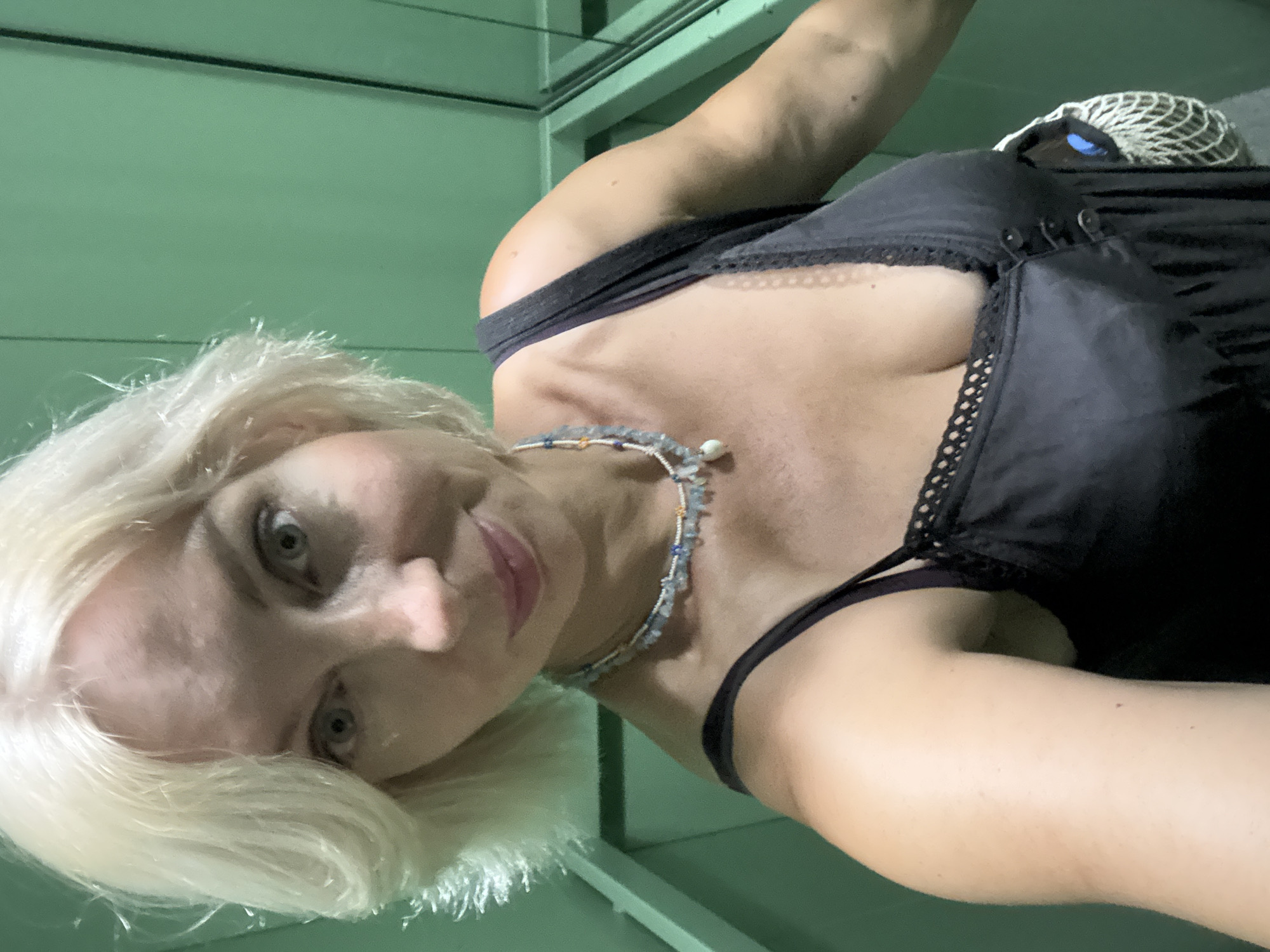A blonde woman in a black dress stands in an elevator with green walls. She is wearing a necklace with blue and white beads and a white pearl pendant. She is holding a white net bag with a blue object inside. The elevator appears to be located in the city of Sochi, Russia.