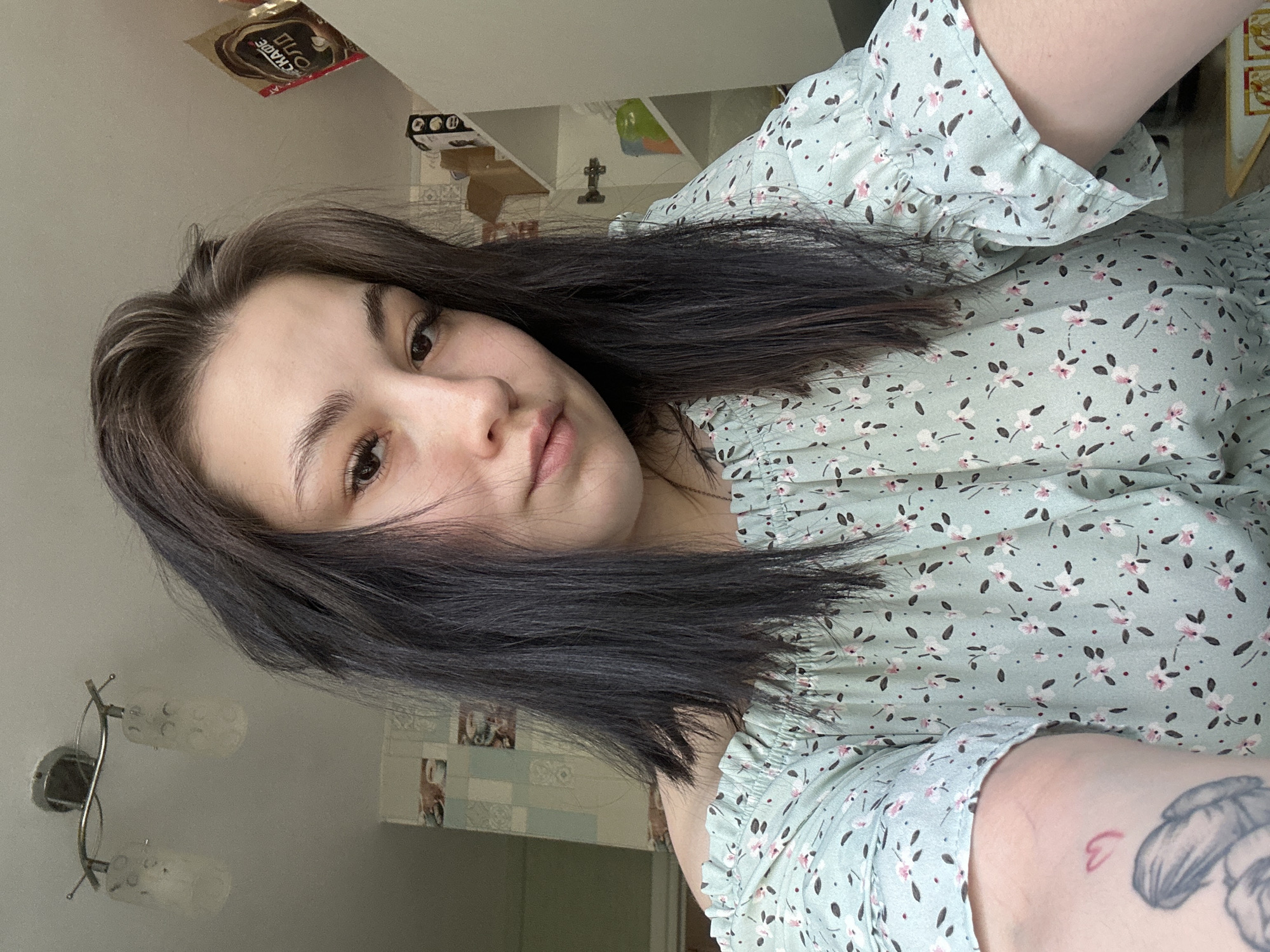 A selfie portrait of a young woman with long, dark hair. She has fair skin and dark eyes. She is wearing a light green floral print dress with ruffled sleeves. The background is a residential interior, including a wall-mounted light fixture, tiled wall accents, shelves with items on them, and a package of coffee. The image was likely taken indoors. There is no information indicating this image was taken in the city of Chaikovsky, Russia.