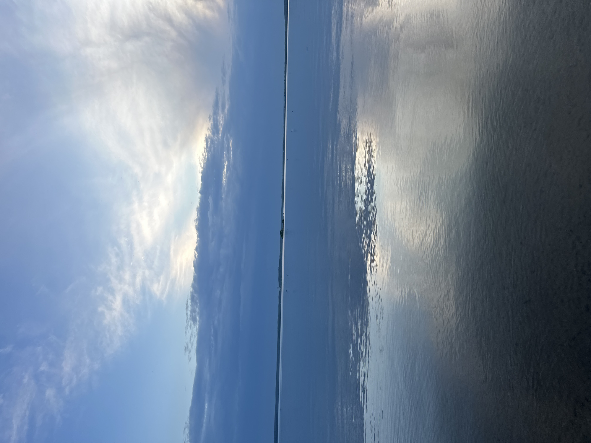 The image shows a body of water, likely a lake or wide river, under a sky with varying cloud cover. The sky on the left is bright blue with wispy clouds, transitioning to a line of heavier, darker clouds toward the center. This cloud formation is mirrored in the water's reflection on the right side of the image. The water is generally smooth, but with subtle ripples creating texture and reflecting the sky. There's a thin line of land visible on the horizon in the center of the image. The exact location in Ждановіцкі сельскі Савет, Belarus, cannot be determined solely from the visual information.