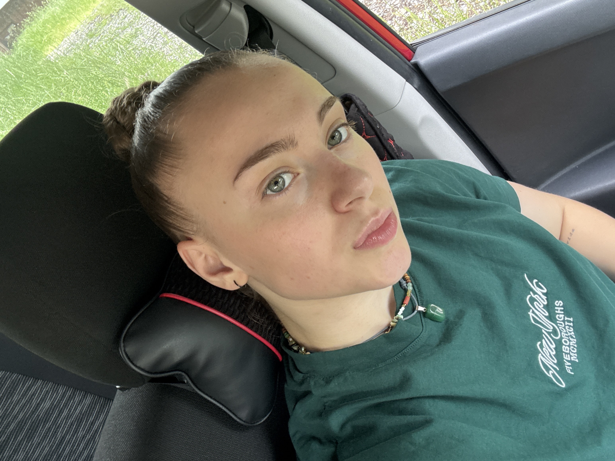 The media file is a close-up selfie taken by a young woman lying down, possibly in a car. She has light skin, green eyes, and her light brown hair is pulled up into two buns on top of her head. She is wearing a dark green t-shirt with white text that appears to say "New York Five Boroughs MCMXCII". She is also wearing a beaded necklace with a green pendant. A black pillow with red trim is visible behind her head. The background suggests an outdoor setting with green grass and some light filtering through. The lighting indicates it is daytime, likely sunny. The woman's expression is neutral, and the overall mood is relaxed. The context provided states the location is Gabrovo, Bulgaria.