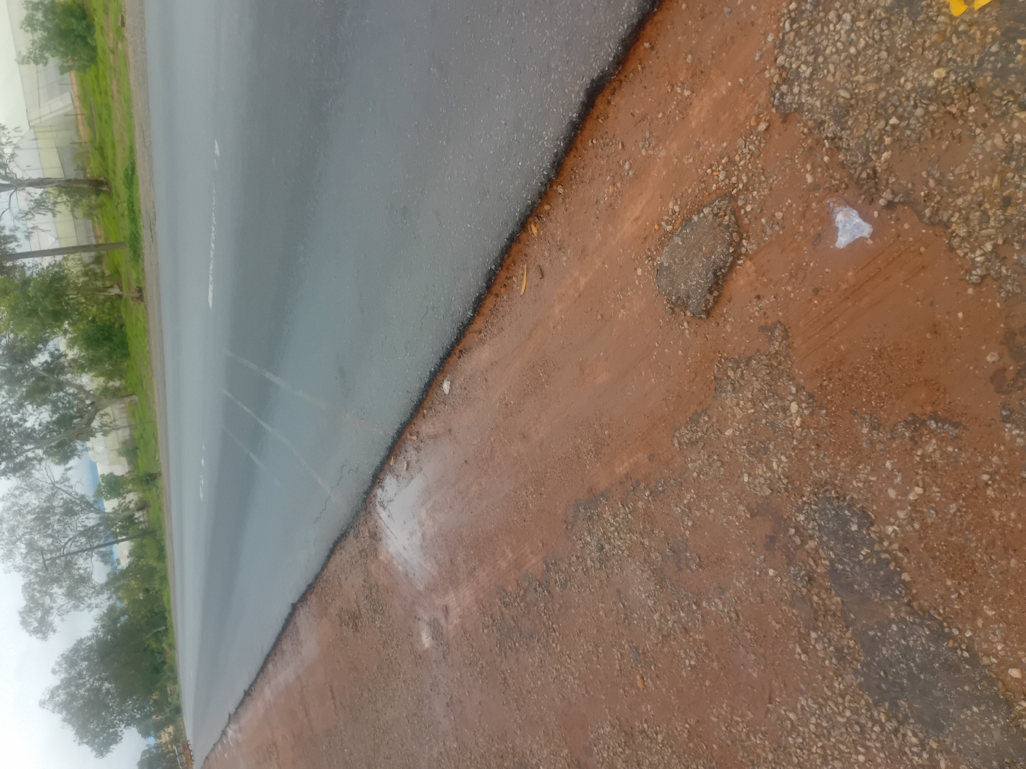 The image depicts a section of a paved road in Jalingo, Nigeria, bordered by a dirt shoulder. The road surface appears to be asphalt, with some faint tire marks visible. The shoulder is composed of reddish-brown dirt mixed with gravel and small stones. There are puddles of water on the shoulder, suggesting recent rainfall or a drainage issue. Some debris, including a white plastic bag and a piece of broken asphalt, is scattered on the ground. In the background, trees line the side of the road, and a low-rise building with a white, possibly corrugated metal roof can be seen in the distance, partially obscured by foliage. The sky is overcast, contributing to a subdued lighting condition. The overall scene suggests a somewhat neglected or under-maintained roadside environment.