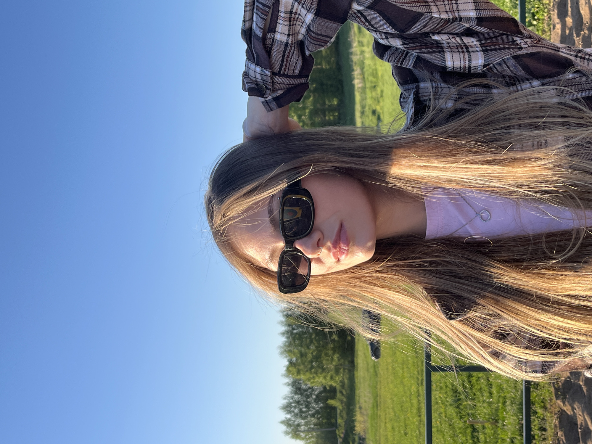 A young woman is posing outdoors on a sunny day. She is wearing sunglasses, a light purple t-shirt, and a brown and white plaid shirt. Her long hair is blond and slightly windswept. She has her right hand raised to the back of her head. In the background, there is a field of green grass with trees and a clear blue sky. It is not possible to ascertain if the photo was taken in the city of Фоки, Russia.