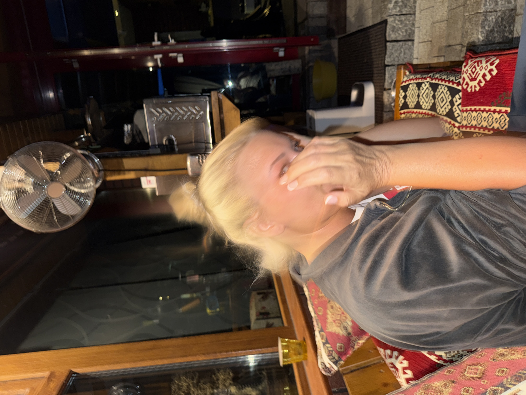 A medium shot captures a blonde woman in the foreground, her face partially obscured by her hands. She is seated on a bench with a richly patterned cushion. She wears a dark grey velour jacket over a white shirt. Behind her, a large window reflects a dark, indistinct interior. Above her head, a metal fan is mounted to the ceiling. To the right, a red-framed doorway leads to an outdoor area with stone steps. A yellow bucket sits on the ground near the doorway. The scene appears to be an indoor seating area, possibly a cafe or restaurant, with elements suggesting it is located in Heiligenrode, Germany, though specific landmarks are not visible.