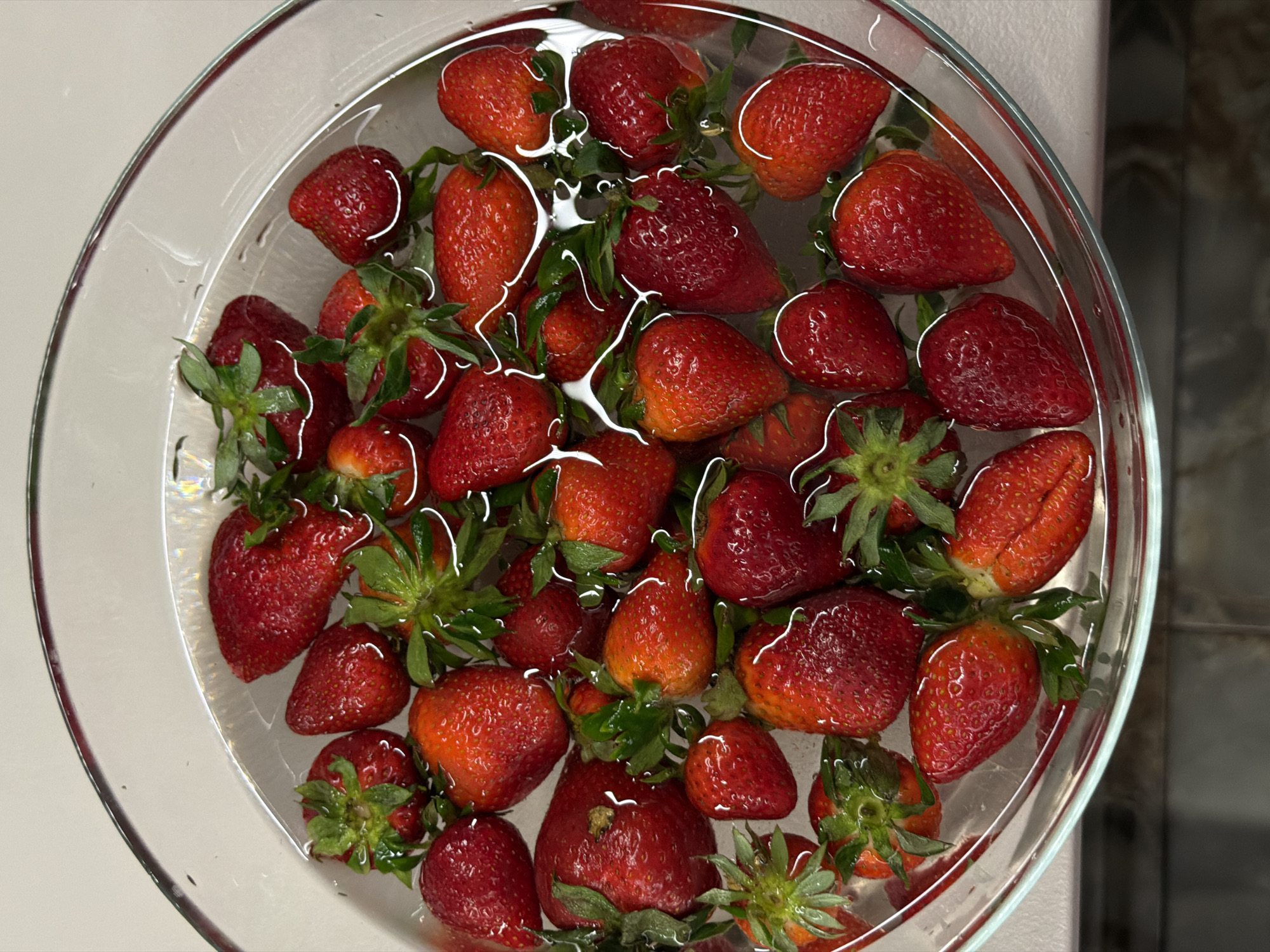 A close-up, top-down shot captures a clear glass bowl filled with numerous red strawberries soaking in water. The bowl is round and transparent, allowing the viewer to see the water and fruit clearly. Most of the strawberries are ripe, displaying vibrant shades of red, though a few show lighter, less ripe areas or tips. Many still have their green calyxes (stems and leaves) attached, which are partially submerged in the water.
The water in the bowl is clear, and its surface reflects ambient light, creating bright, wavy lines and patterns across the strawberries. This suggests movement or ripples in the water, or perhaps just the effect of light hitting the surface. The strawberries vary in size, with some appearing large and plump, and others smaller.
The background on the left and top of the image is a light, uniform surface, likely a countertop or table. To the right, the background darkens and shows a subtle texture, possibly indicating a wall or kitchen cabinets. The lighting is bright and even, highlighting the fresh appearance of the fruit. No people are visible, and there is no discernible text. The scene depicts a simple, everyday act of preparing fresh fruit, possibly in a kitchen in Moscow, Russia.