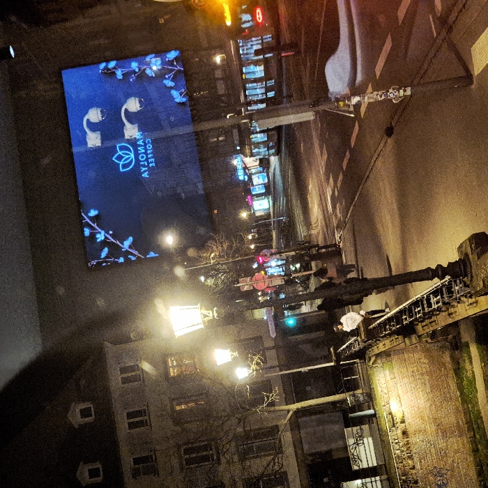 A nighttime street scene with a modern advertisement displaying "AYTOMAT COFFEE" alongside images of wireless earbuds and floral designs. A series of street lights illuminate the road, reflecting off the wet pavement. Pedestrians walk along the sidewalk. A brick wall with graffiti is visible in the foreground, contrasting with the modern advertising. A multi-story building stands across the street, casting long shadows in the dark.