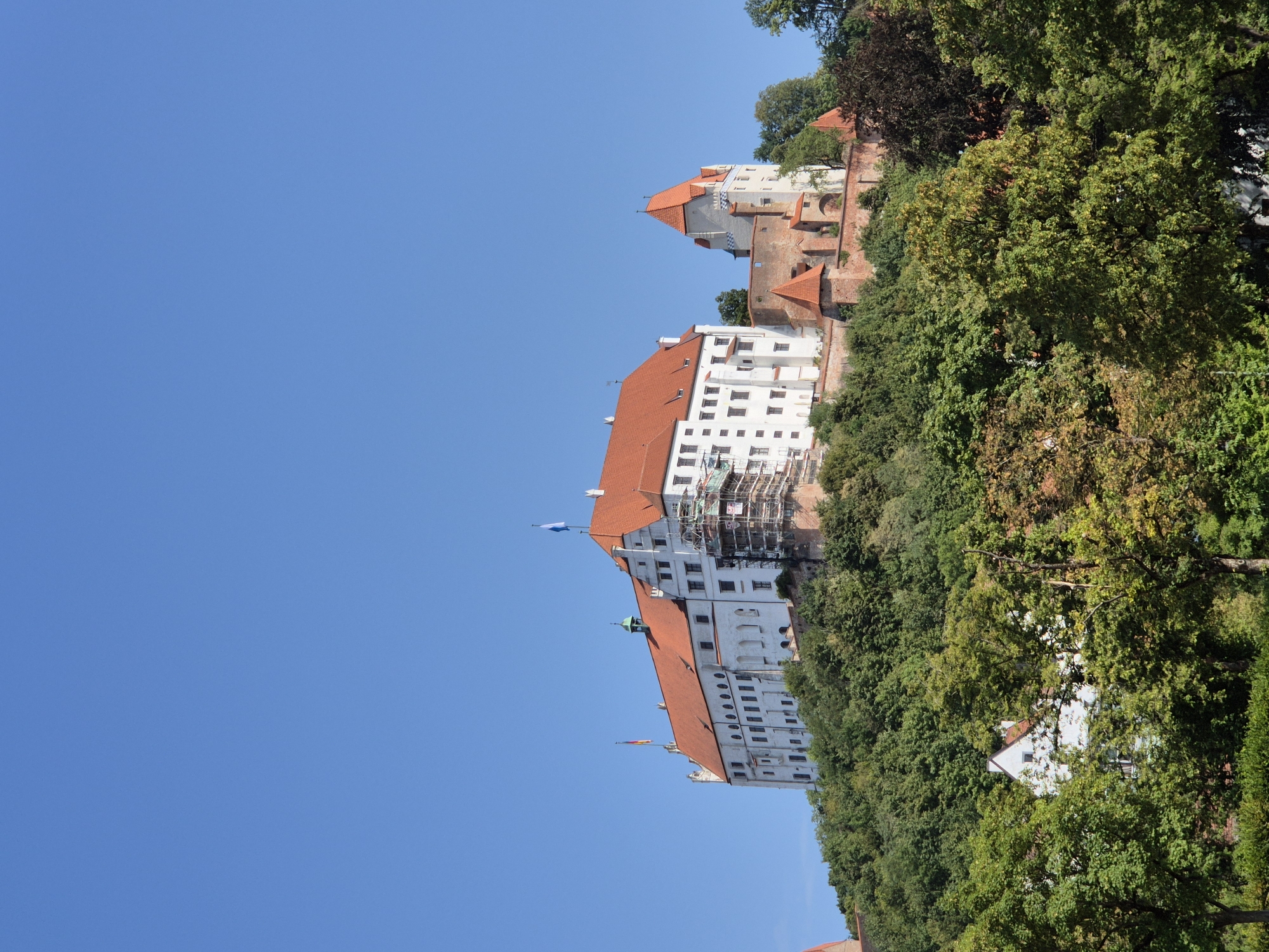 The image shows Trausnitz Castle, located on a hill overlooking the city of Landshut, Germany. The castle is a large, white building with a red tile roof, and it is surrounded by trees. The sky is clear and blue. There are flags on top of the castle. Scaffolding is visible on one side of the castle, indicating construction or maintenance work. The castle is a prominent landmark in Landshut and is a popular tourist attraction.