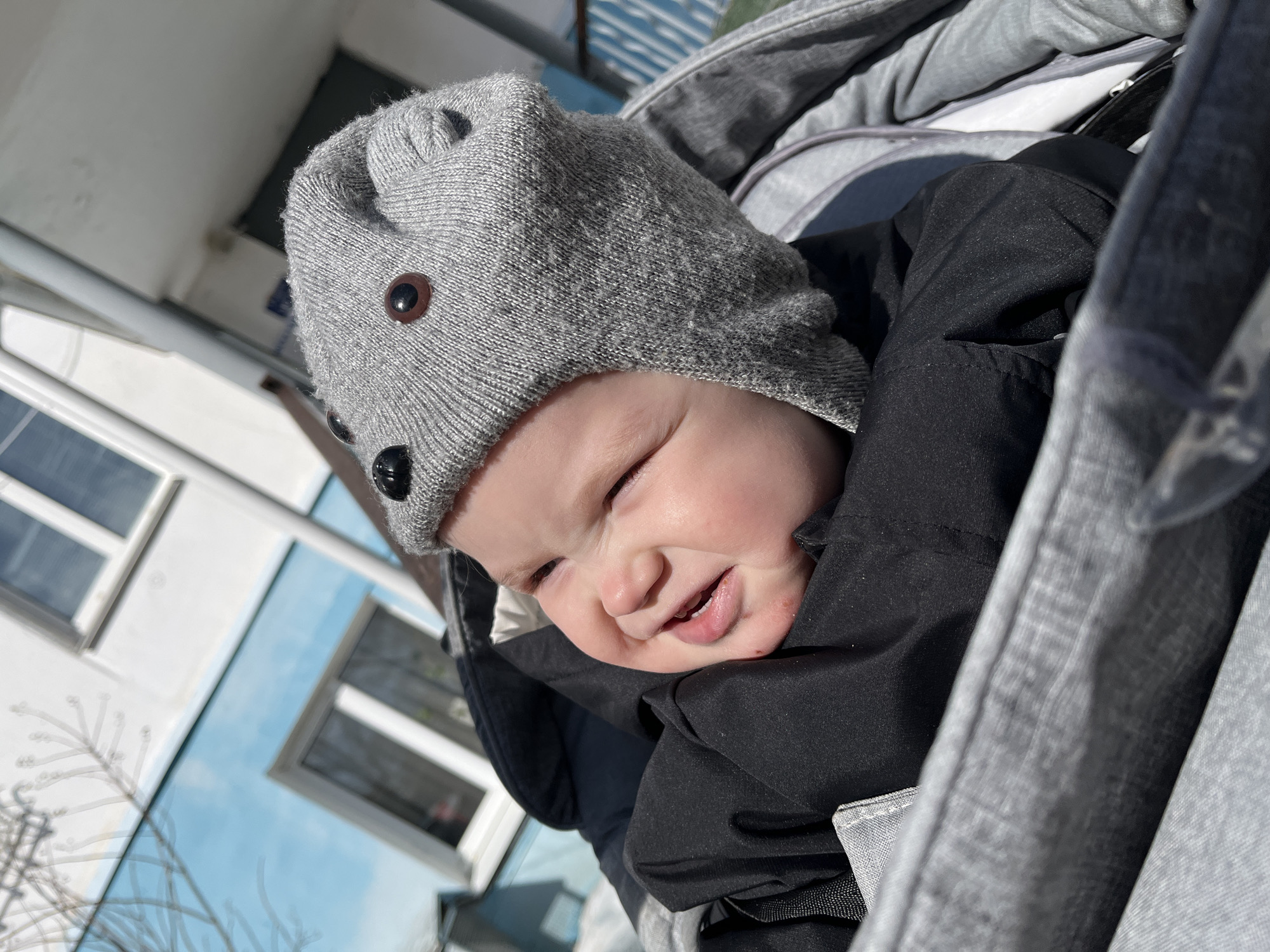 A baby is positioned in a grey fabric stroller or carrier, centered in the frame. The baby, with fair skin, wears a grey knitted hat featuring two dark brown circular buttons resembling eyes and two black circular buttons for a nose. A dark grey or black jacket covers the baby's upper body. The baby's mouth is open, exposing gums, and the eyes are squinted, suggesting a smile or reaction to bright sunlight. A small reddish mark is present on the lower right cheek. The background features a multi-story building, with the lower section painted light blue and the upper section white. Rectangular windows with white frames are visible on the building. Bare tree branches are partially visible in the upper left background. The scene is brightly illuminated by direct sunlight, casting shadows. The specific location within Chaikovsky, Russia, is not identifiable from the visual evidence.