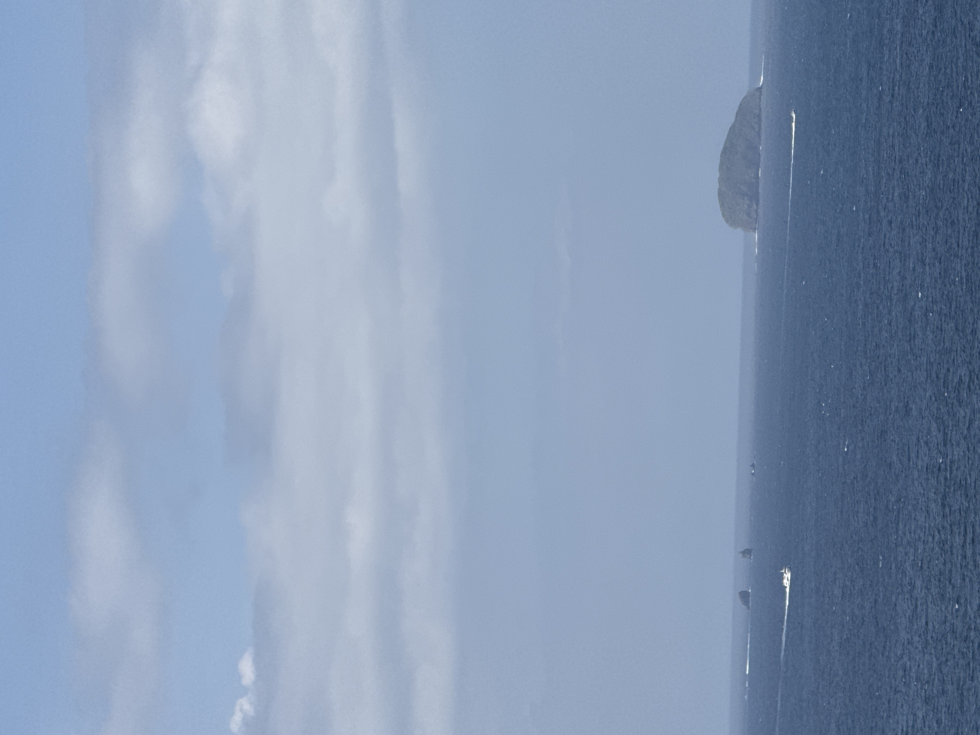 A wide shot captures a calm, deep blue ocean under a sky filled with pale, wispy clouds. In the distance, a small, rounded, vegetated island sits on the horizon. Several boats are visible on the water: one is prominently in the foreground, leaving a white wake as it speeds across the sea. Further back, other boats are dotted across the expanse, some near small rock formations or other distant islands. The scene is serene and expansive, suggesting an open ocean environment.