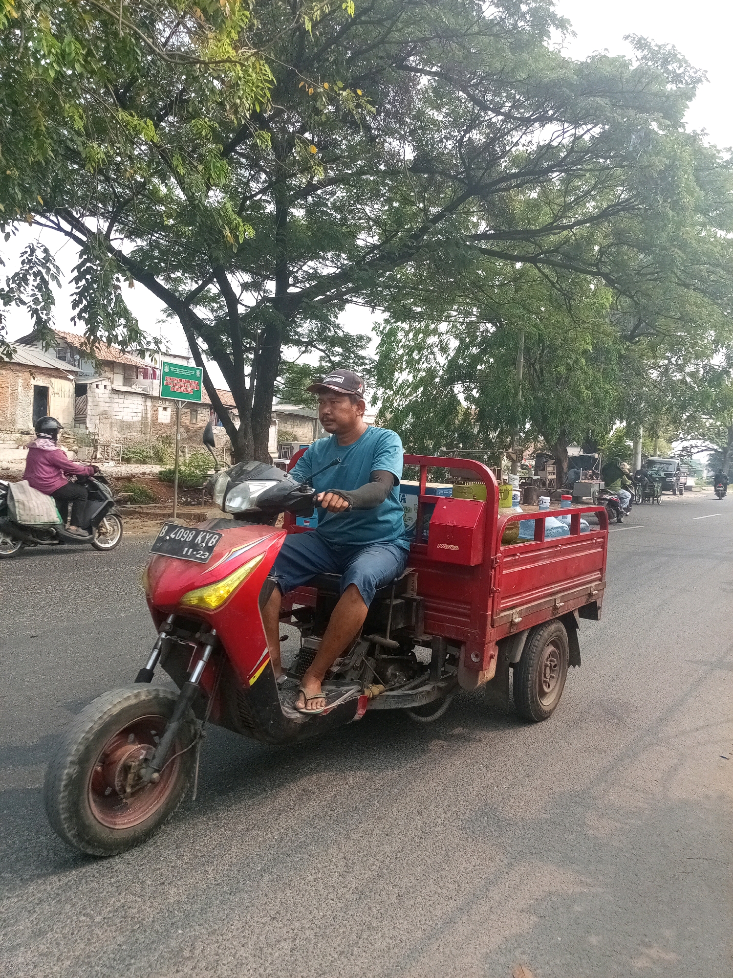 A man wearing a blue t-shirt, blue shorts, a baseball cap, and sandals rides a red three-wheeled scooter with a cargo bed on a paved road. He is wearing arm sleeves. In the cargo bed are several large water bottles and other items. The scooter has a license plate that reads "B 4098 KYB 11-23". The scooter is traveling on the right side of the road. In the background, there are buildings, trees, and other vehicles including a scooter with a rider wearing a pink shirt and black helmet. There is also a green sign visible with Indonesian text. The scene appears to be in the city of Bekasi, Indonesia, indicated by the license plate format and the environment.