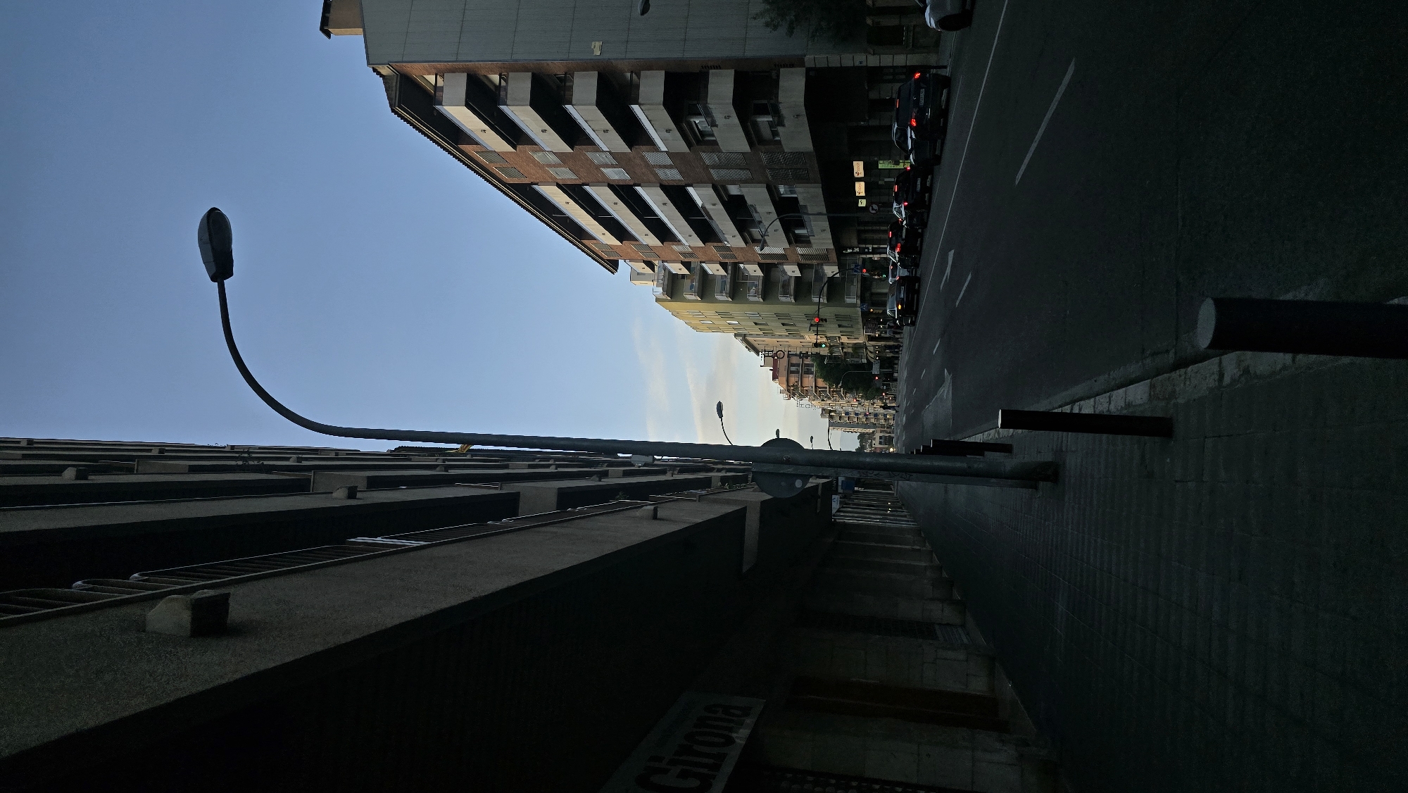 This image captures an urban street scene from a low, tilted perspective, looking upwards along a thoroughfare lined with buildings. The composition creates a dynamic and somewhat disorienting view, as if the camera were angled sharply towards the sky and rotated.
On the left side of the frame, a tall, dark building with prominent, shadowed balconies or ledges dominates the foreground. Emerging from its base and curving gracefully upwards across the clear, light blue sky is a slender streetlamp with a distinctive, elongated head, silhouetted against the bright backdrop.
On the right side, a paved street with faint white lane markings runs parallel to a row of buildings. Several parked cars are visible along the street, with some rear lights reflecting light. Beyond these, a succession of multi-story buildings with varying facades – some brick, others in lighter tones like yellow or pale green – recedes into the distance. Small traffic lights can be discerned further down the street. The sidewalk alongside the street features several dark, cylindrical and rectangular posts or bollards.
The lighting conditions suggest either early morning or late evening. The upper portions of the buildings catch some ambient light, while the street and lower sections are cast in deep shadow, creating high contrast. The sky is clear, indicating fine weather. Partially visible on a sign or poster on the dark building in the foreground is the word "Girona." No overt activity is taking place, contributing to a sense of quiet.