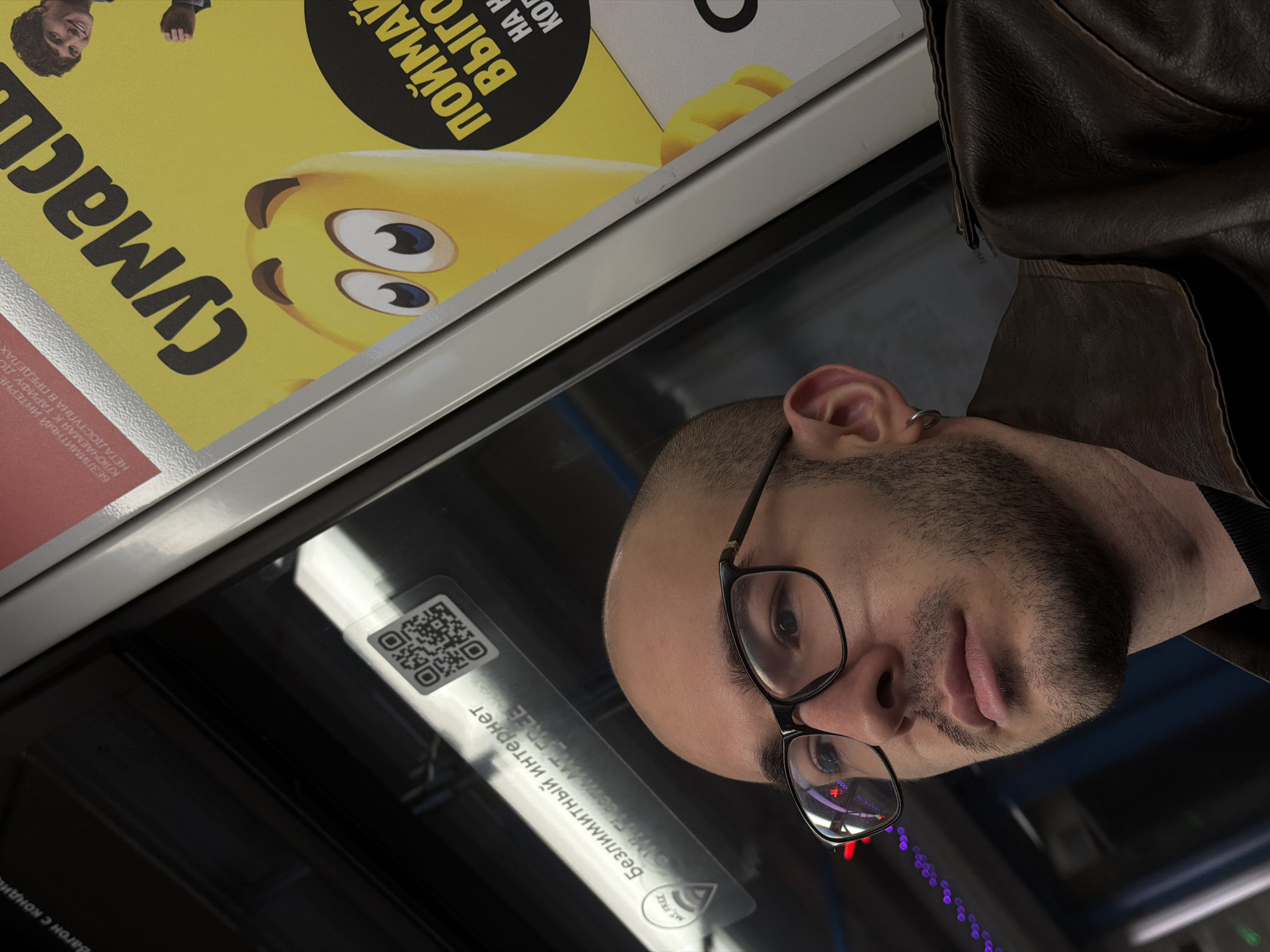 A bald man with glasses and a brown leather jacket stands in the Moscow metro. He is looking directly at the camera. In the background is an advertisement that reads "Сумас." and a sticker that says "Бесплатный интернет" (Free internet). There is also a QR code visible.