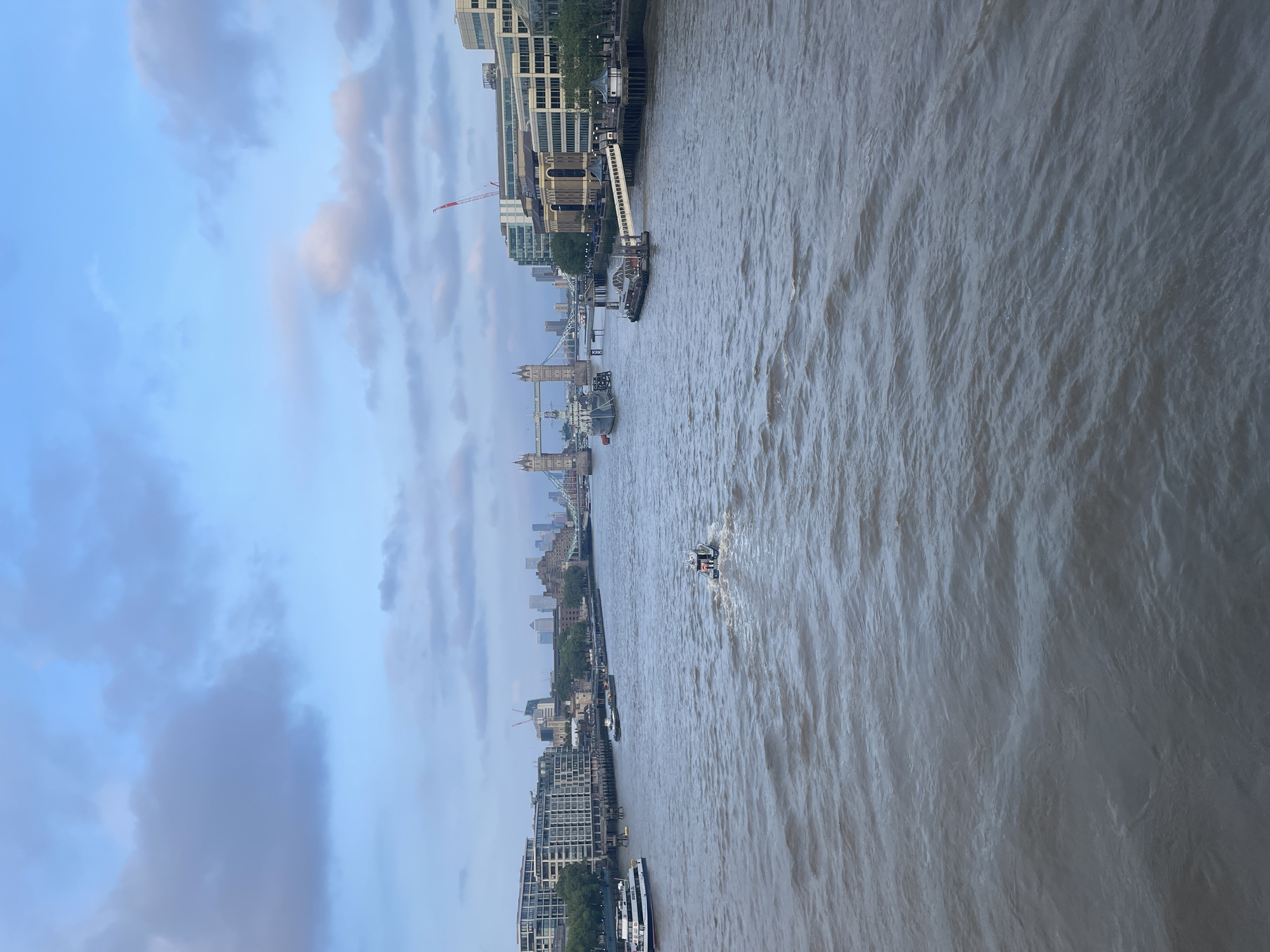 The image shows a view of the River Thames in London, United Kingdom. The iconic Tower Bridge is visible in the mid-ground, connecting two banks. Tall buildings line the riverbanks, indicating a densely built urban environment. A boat is on the water, creating a wake. The sky is cloudy with a mix of blue and grey. A crane can be seen amongst the buildings along the river. The river appears choppy, indicating movement of water.