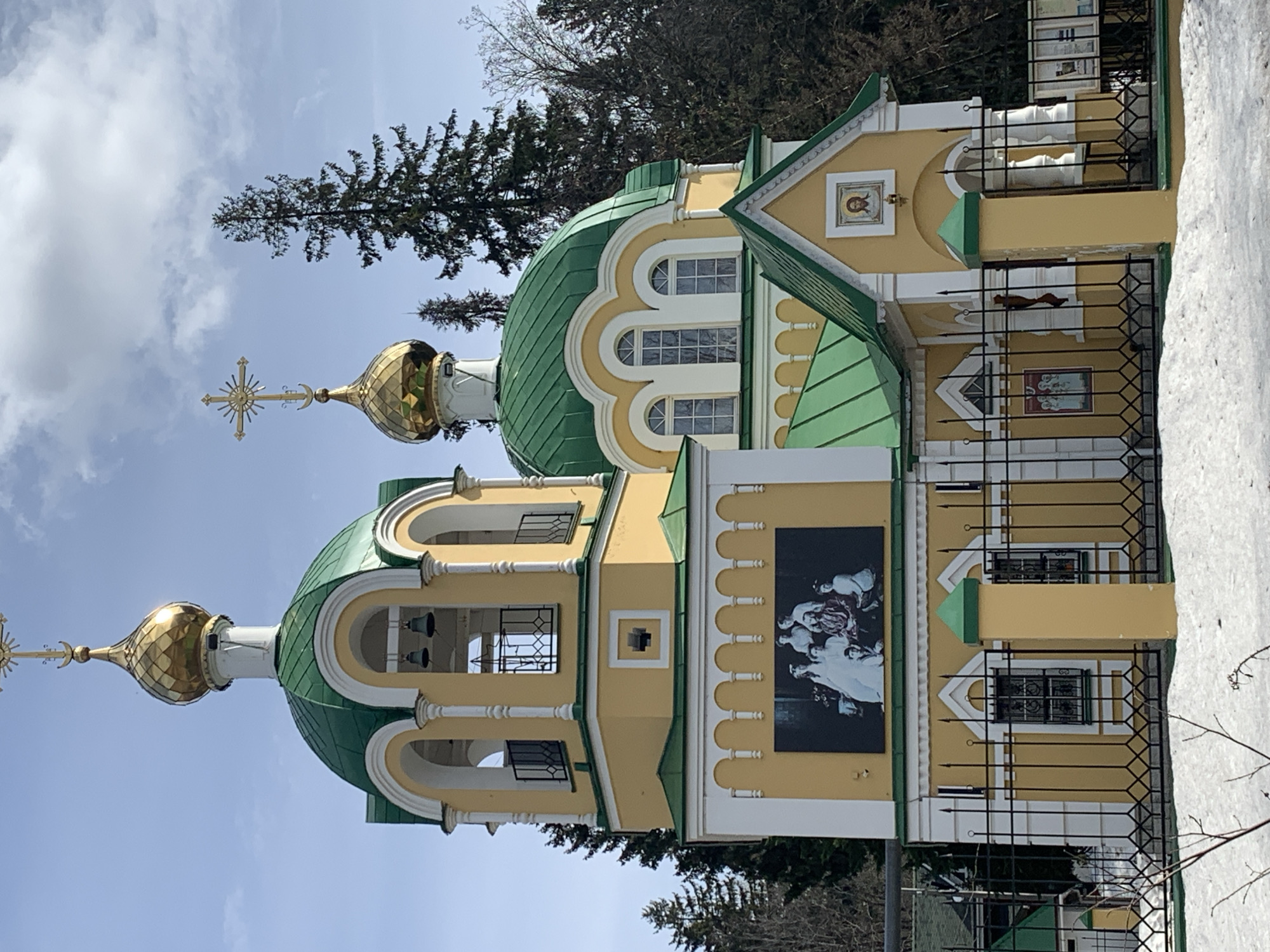 The image shows a yellow Orthodox church building with green roofs and gold domes in Izhevsk, Russia. The building has decorative arched windows and a large black and white mural. A black metal fence borders the building, and patches of snow are visible in the background. Trees are seen behind the church, and the sky is visible above.