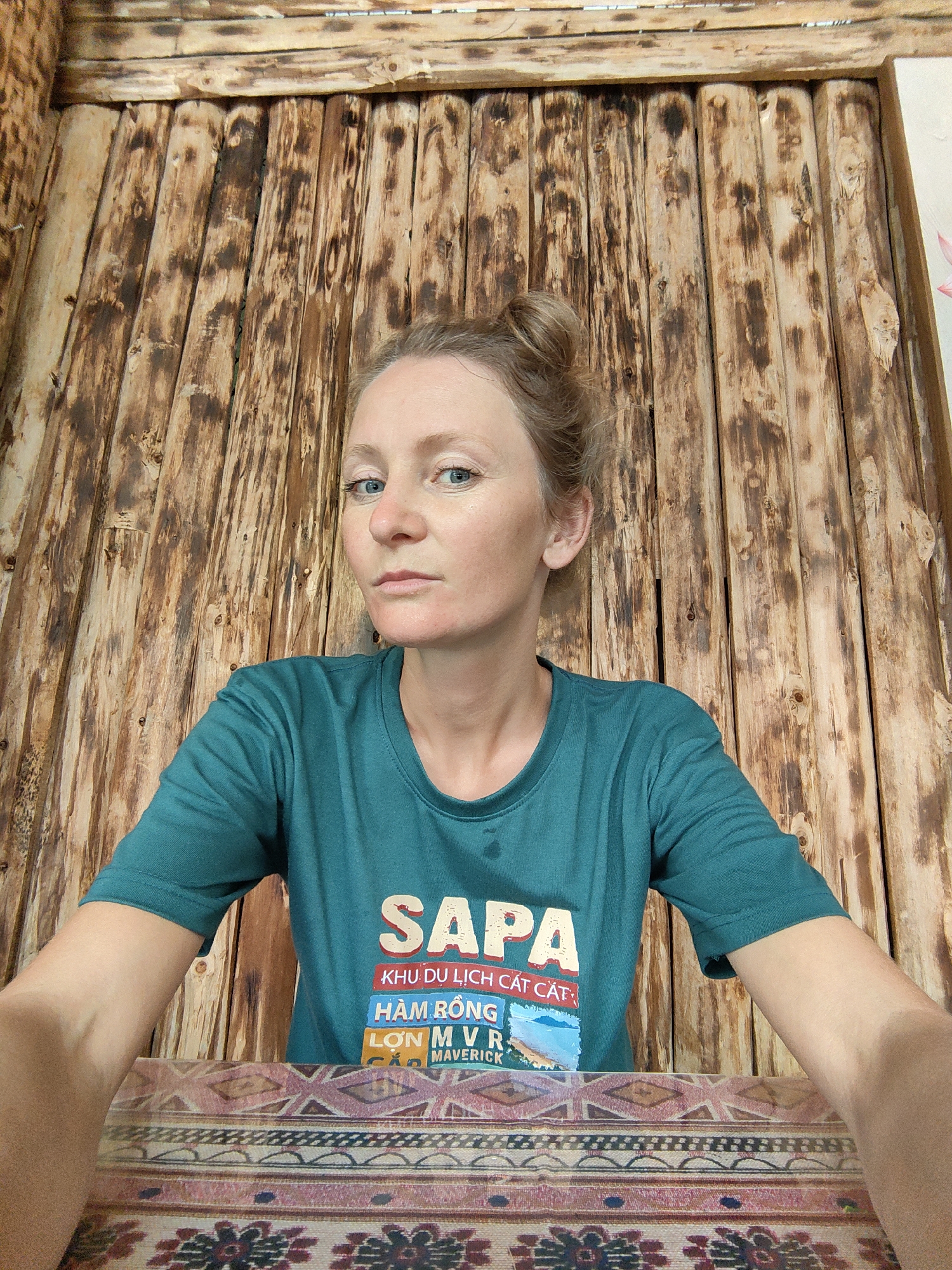 A medium shot captures a fair-skinned woman with blonde hair styled in a bun. She is positioned indoors, wearing a teal t-shirt with "SAPA" printed in large white letters above smaller text in Vietnamese and English, including "KHU DU LỊCH CÁT CÁT," "HÀM RỒNG," "LỚN MVR," and "MAVERICK." The background consists of vertical wooden planks with a rustic appearance. The location is identified as Thành phố Hà Giang, Vietnam.