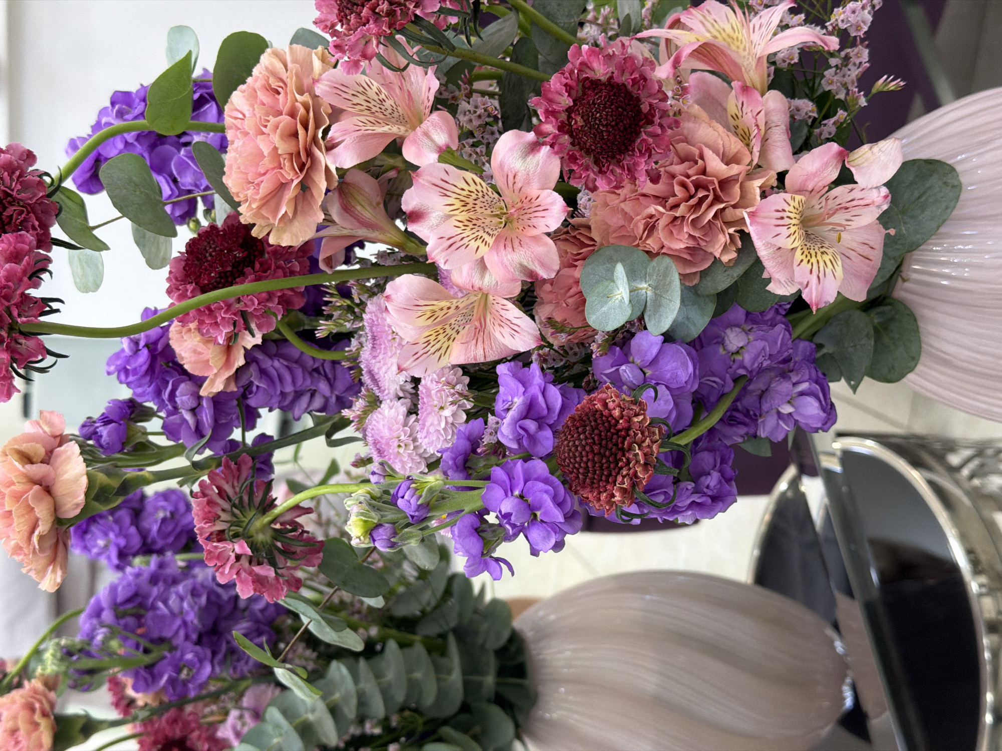 The image depicts an arrangement of flowers, predominantly in shades of pink and purple, with some green foliage. The types of flowers appear to include carnations, alstroemeria (Peruvian lilies), and stocks (Matthiola incana). The arrangement is in a light-colored vase with a ribbed texture. In the background, part of a shiny, possibly metal, object is visible. While the image provides no information on the location or the city of Moscow, Russia, it is simply a close-up shot of a floral arrangement.