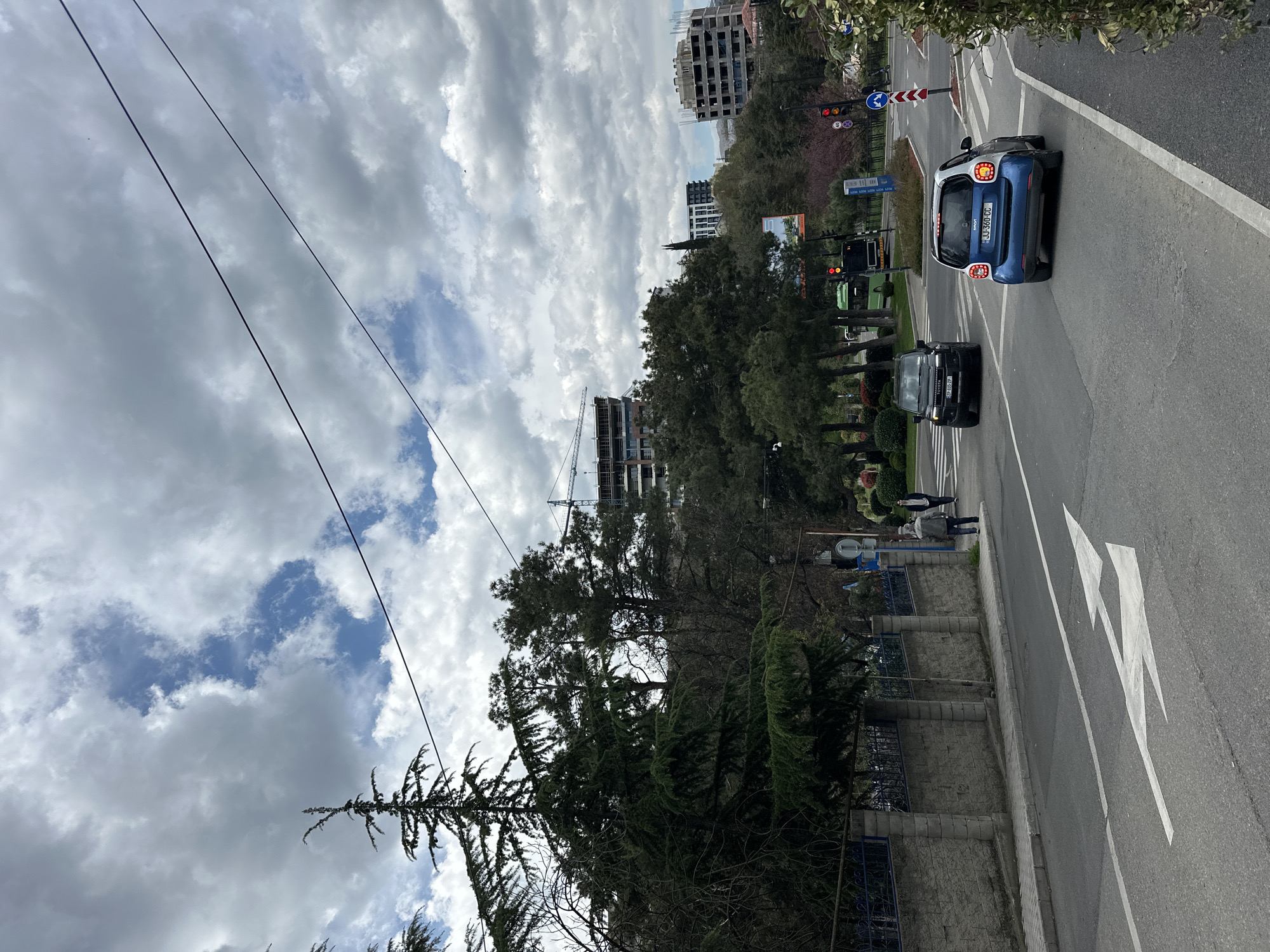 This outdoor photograph captures a bustling urban street scene under a cloudy sky with visible patches of blue. Thin power lines stretch across the upper part of the frame.
The foreground features a road with distinct white markings, including a large white arrow painted on the asphalt, indicating a turn. Two cars are prominent on the road: a small blue and white Smart car, displaying the license plate "JJ-380-CC", and a black SUV with the license plate "TP-007P" positioned behind it. Further down the road, a green bus is partially visible. Traffic lights, with one showing red, and various road signs—including a blue circular mandatory direction sign and a red and white chevron sign—are visible at an intersection.
To the right of the road, a sidewalk is present, bordered by a low stone wall topped with a blue metal fence. Two individuals are walking on this sidewalk. The area is rich with vegetation, featuring numerous tall evergreen trees and other trees lining the street, suggesting a park-like area adjacent to the road.
In the background, several buildings contribute to the urban landscape. One multi-story structure appears to be under construction, marked by a large crane beside it. Other completed buildings are also visible. The overall scene depicts a functional city environment during daytime, characterized by a mix of infrastructure, natural elements, and human activity.