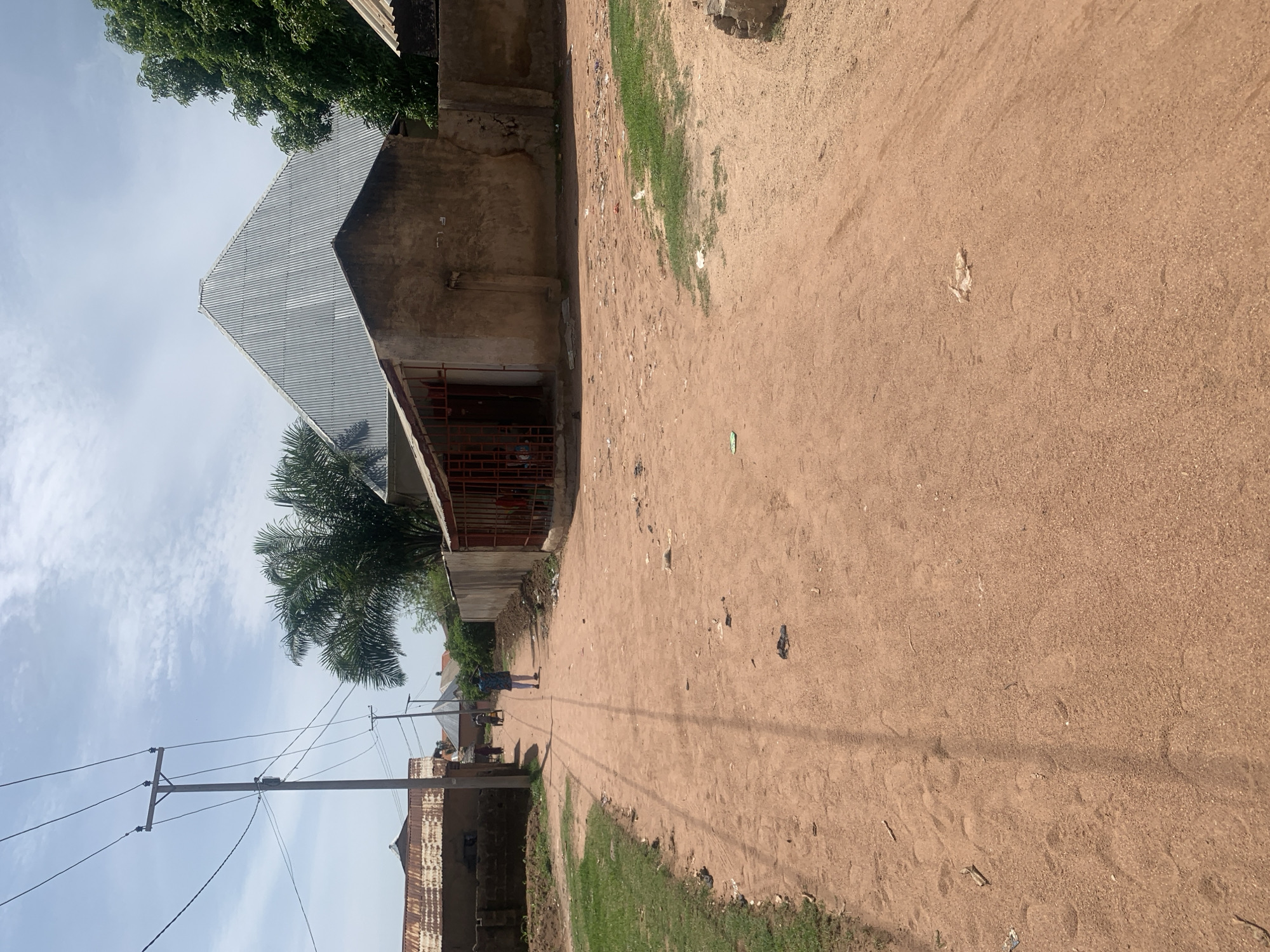 The image depicts a dirt road in a residential area of Jalingo, Nigeria, under a partly cloudy sky. The road is made of reddish-brown earth and shows tire tracks and footprints. On the left side of the road, a grassy area is visible, along with a power pole and overhead electrical wires. Several buildings line the street, featuring corrugated metal roofs and rough-textured walls. One prominent building on the right has a conical, ribbed metal roof. In the middle distance, a few individuals are walking along the road. One person, wearing a patterned shirt and white pants, is facing away from the camera. The environment appears to be a typical, somewhat unkempt, urban or semi-urban setting.
