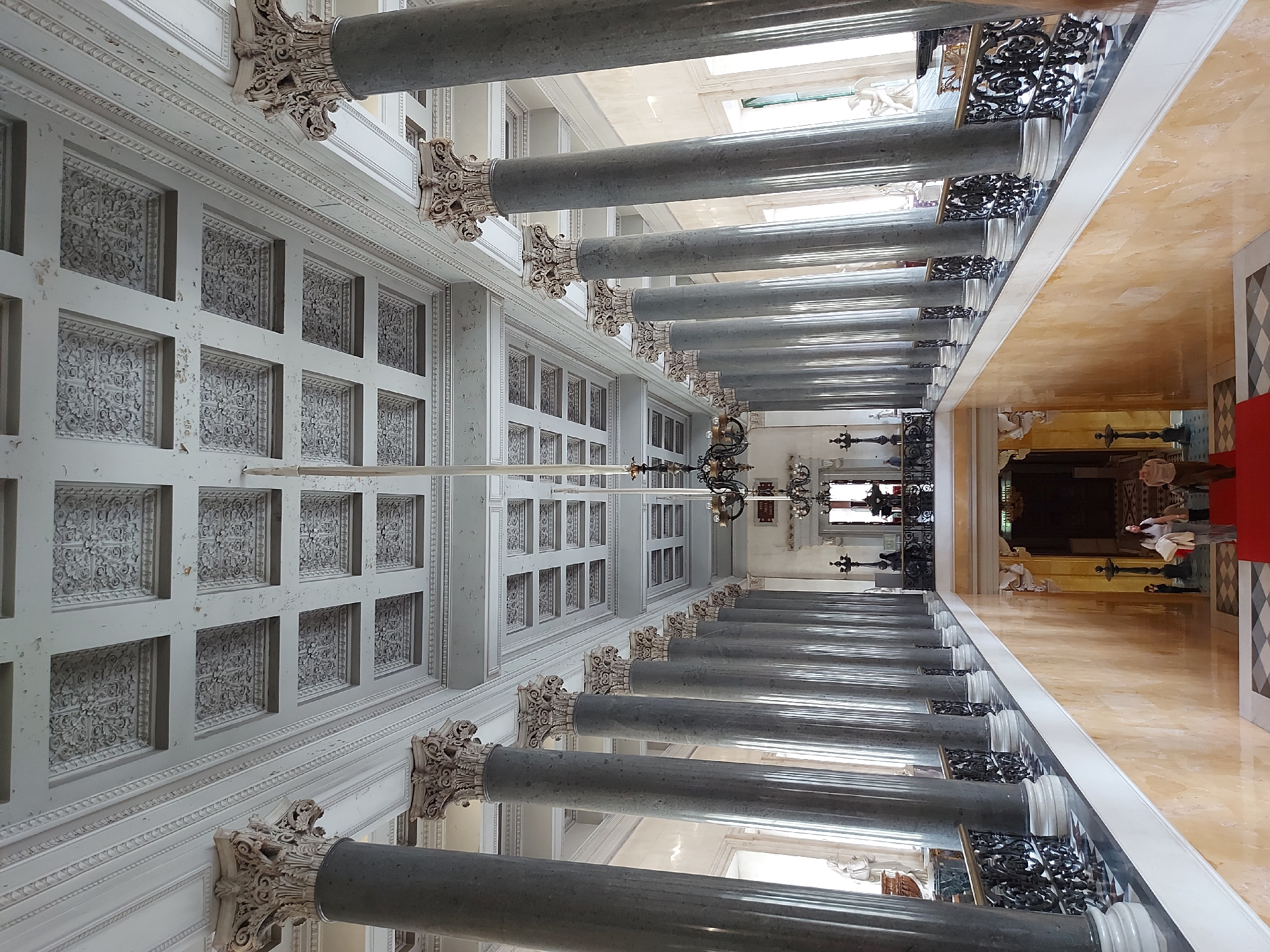The image captures a long hallway in what appears to be an opulent interior, likely a historical building in Saint Petersburg, Russia. The hallway is defined by parallel rows of gray marble columns with ornate, white capitals. Above the columns is a heavily ornamented, coffered ceiling. A long, golden-hued marble floor runs along one side, while the other side features intricate black wrought-iron railing and a white balustrade, topped by white sculptures. Towards the end of the hallway, there is a glimpse of an interior area, possibly a room or chapel, where people can be seen. A red carpet leads into the room. The lighting in the hallway is a mixture of natural light coming from windows above the columns and artificial light from chandeliers hanging from the ceiling. The scene exudes an aura of classical architecture and historical significance.