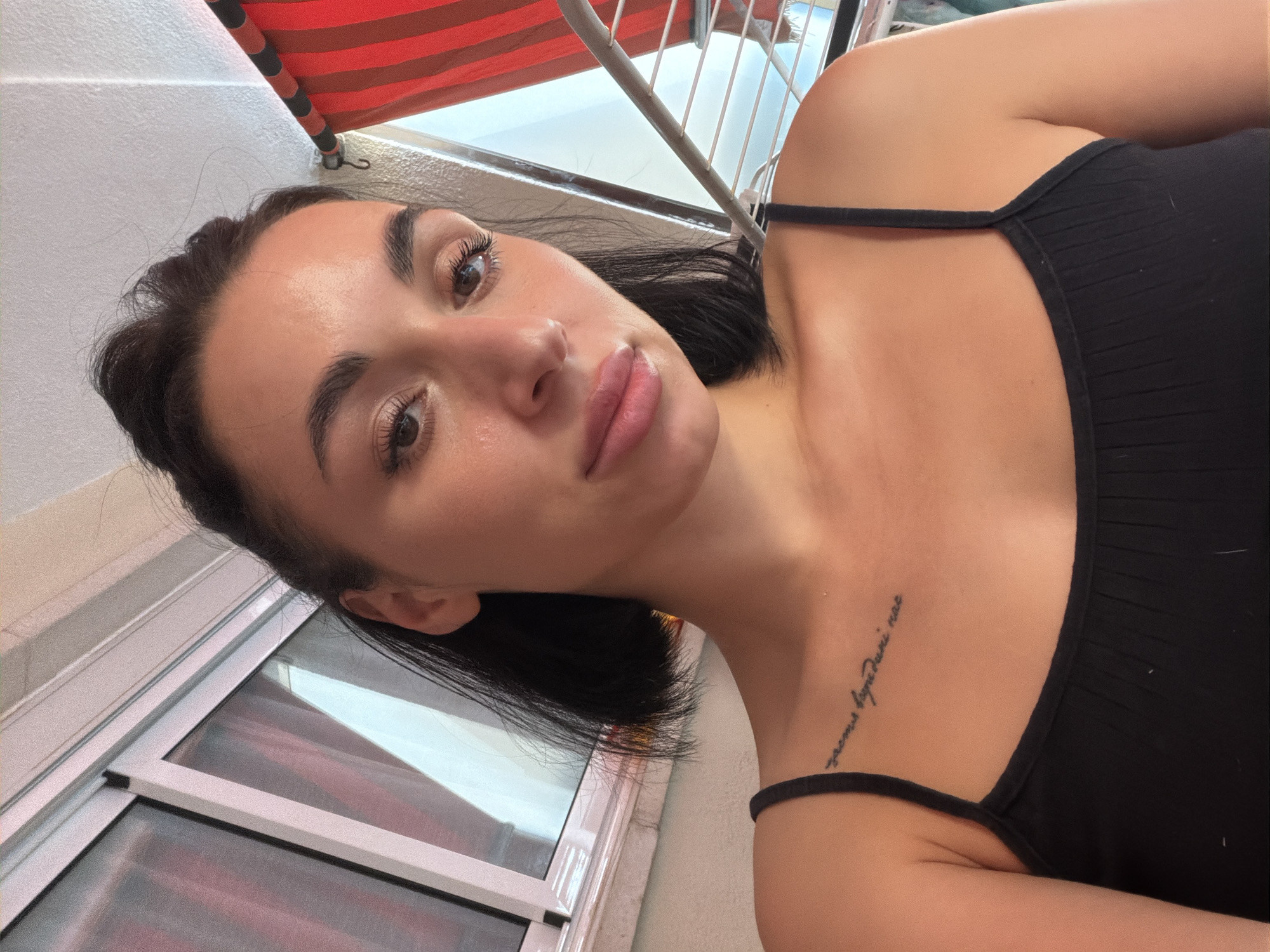 A young woman with dark hair, styled in a bob, is taking a selfie. She is wearing a black camisole. A small tattoo is visible on her upper chest, above her left collarbone. The background shows a window with light pink curtains and a red and white striped awning. There is also a metal drying rack visible. The scene appears to be on a balcony or terrace, as suggested by the awning and the presence of a clothes drying rack, common in outdoor spaces in coastal areas like Salou, Spain. The lighting suggests it is daytime.