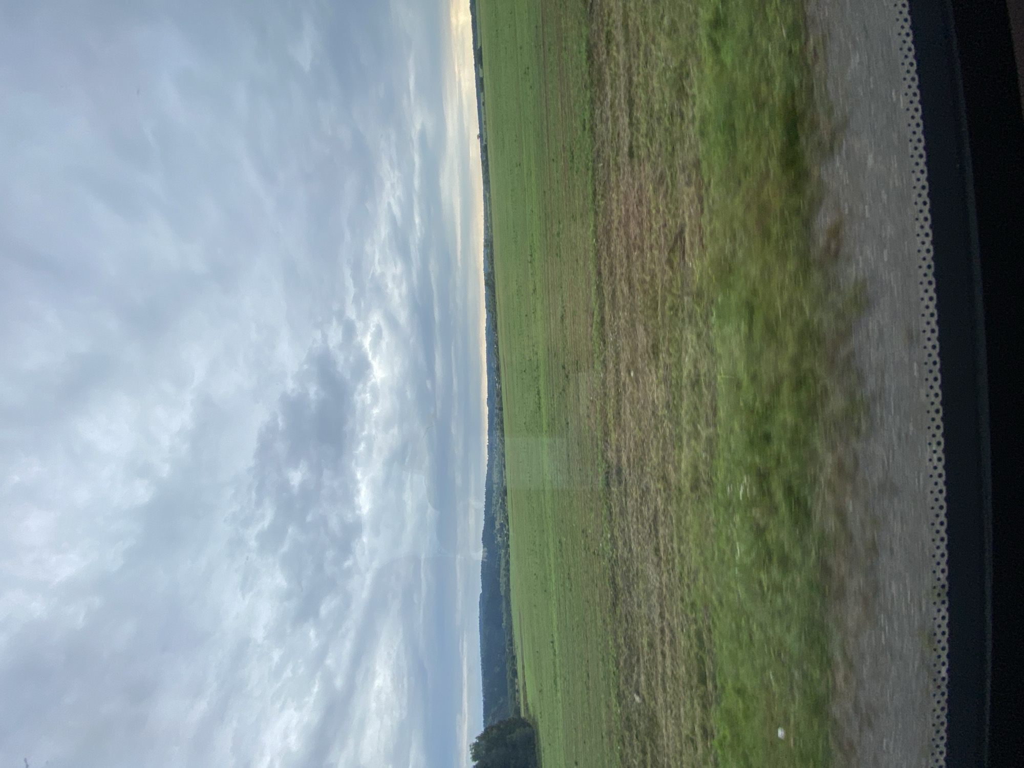 The image depicts a wide, open field under a cloudy sky. The field is predominantly green, with some areas appearing to be recently mown or harvested, showing patches of shorter, dry grass. In the distance, a line of trees and rolling hills forms the horizon, with hints of scattered buildings suggesting a rural settlement. The sky is filled with a mix of grey and white clouds, creating an overcast and somewhat dramatic atmosphere. The foreground shows a blurred view, possibly from a moving vehicle, with a dark surface at the very bottom and a decorative patterned edge along the top of this surface, indicating a window or similar barrier. The scene is devoid of any human presence or activity, focusing solely on the landscape. The location is identified as the city of Bizyar, Russia.